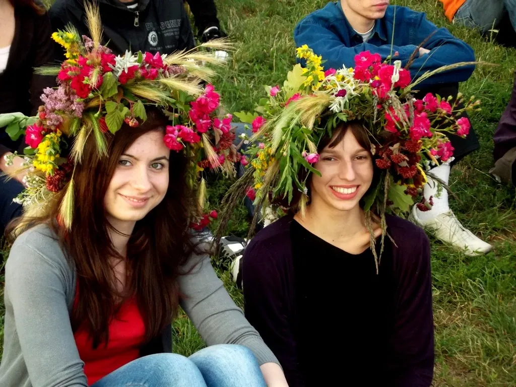 Two young women wearing large, colorful wreaths of flowers and grain sit smiling on the grass.
