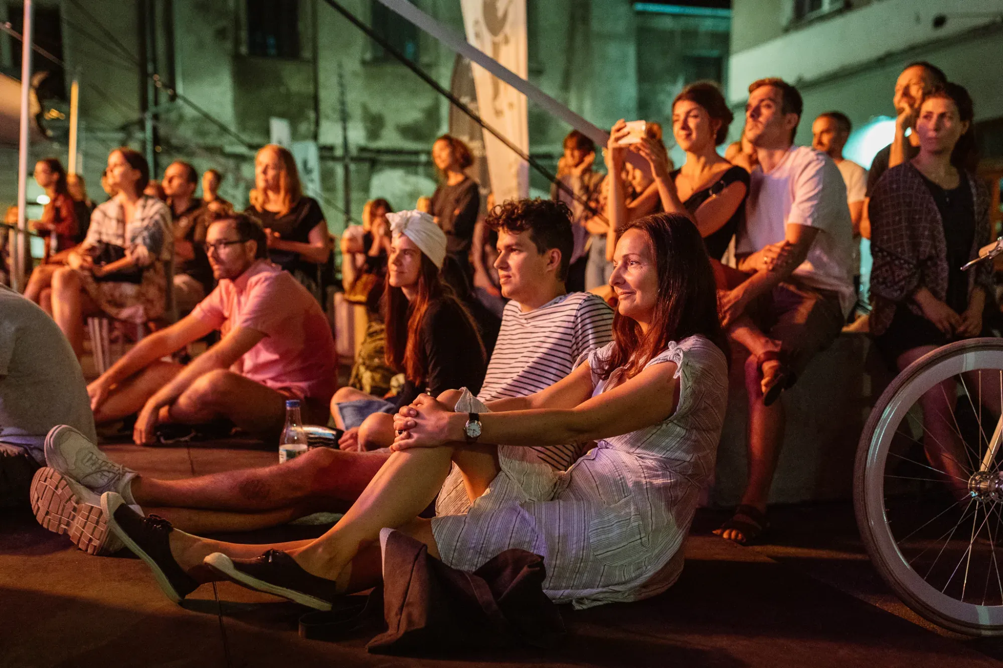 A crowd of young people sits on the ground at night, watching an open-air performance during Jewish Culture Festival