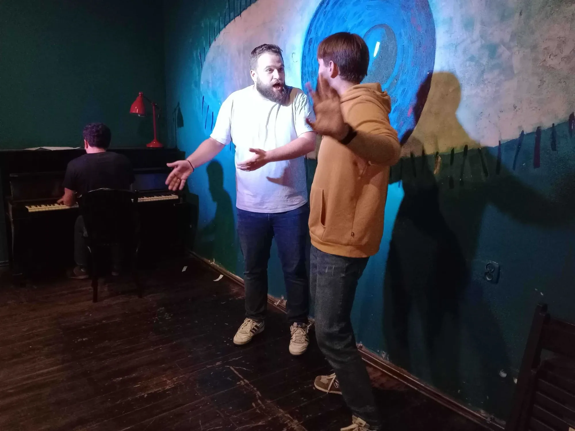 Zwei Männer singen oder spielen enthusiastisch, während im Hintergrund ein Mann Klavier spielt.