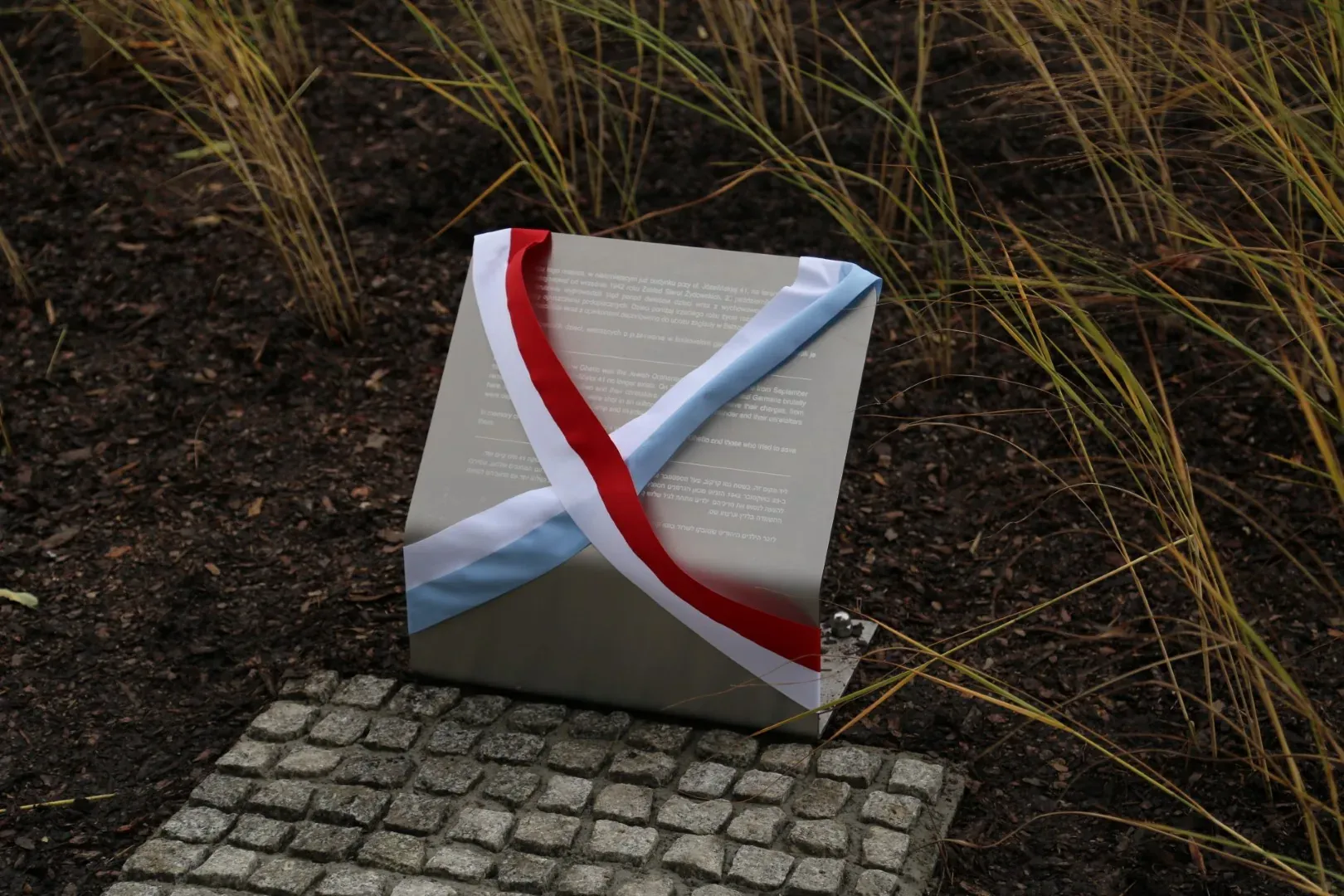 A memorial plaque with red, white, and blue ribbons, set in the ground with cobblestones and tall grass.