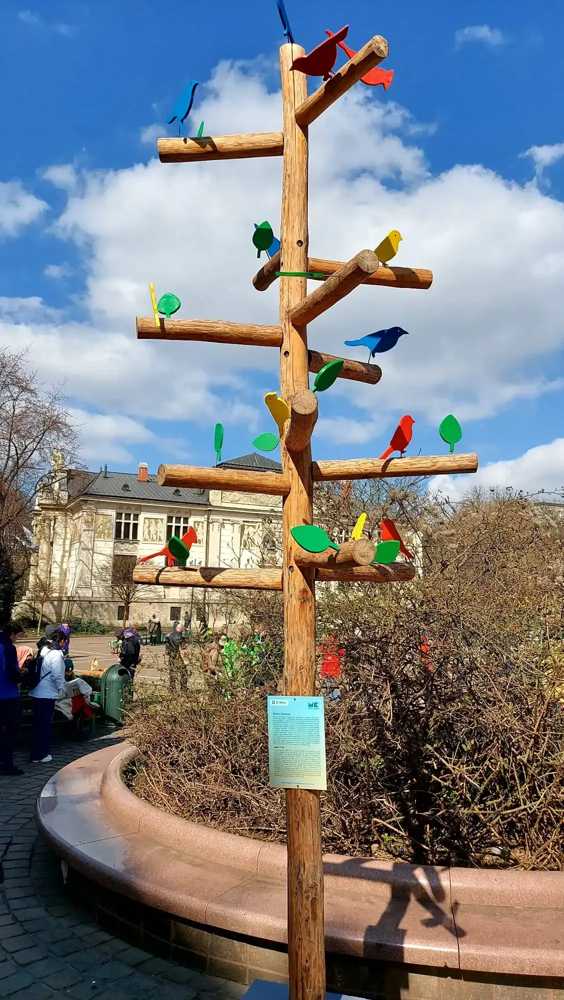 Tall wooden tree-like sculpture with colorful bird and leaf cutouts under a blue sky. People and a historical building in the