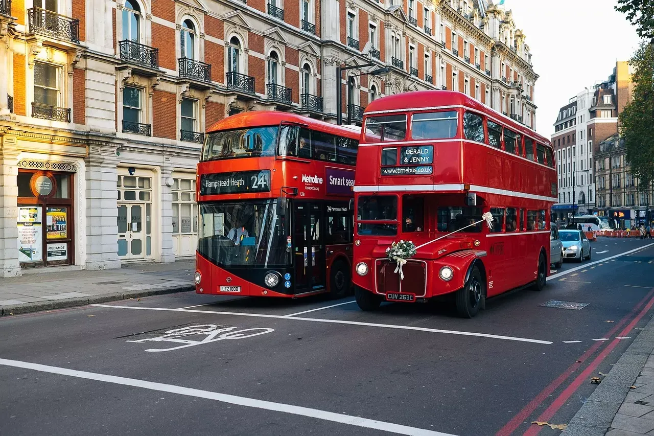 Zwei rote Doppeldeckerbusse auf einer Londoner Straße. Einer ist ein öffentlicher Bus, der andere ein alter Hochzeitsbus mit