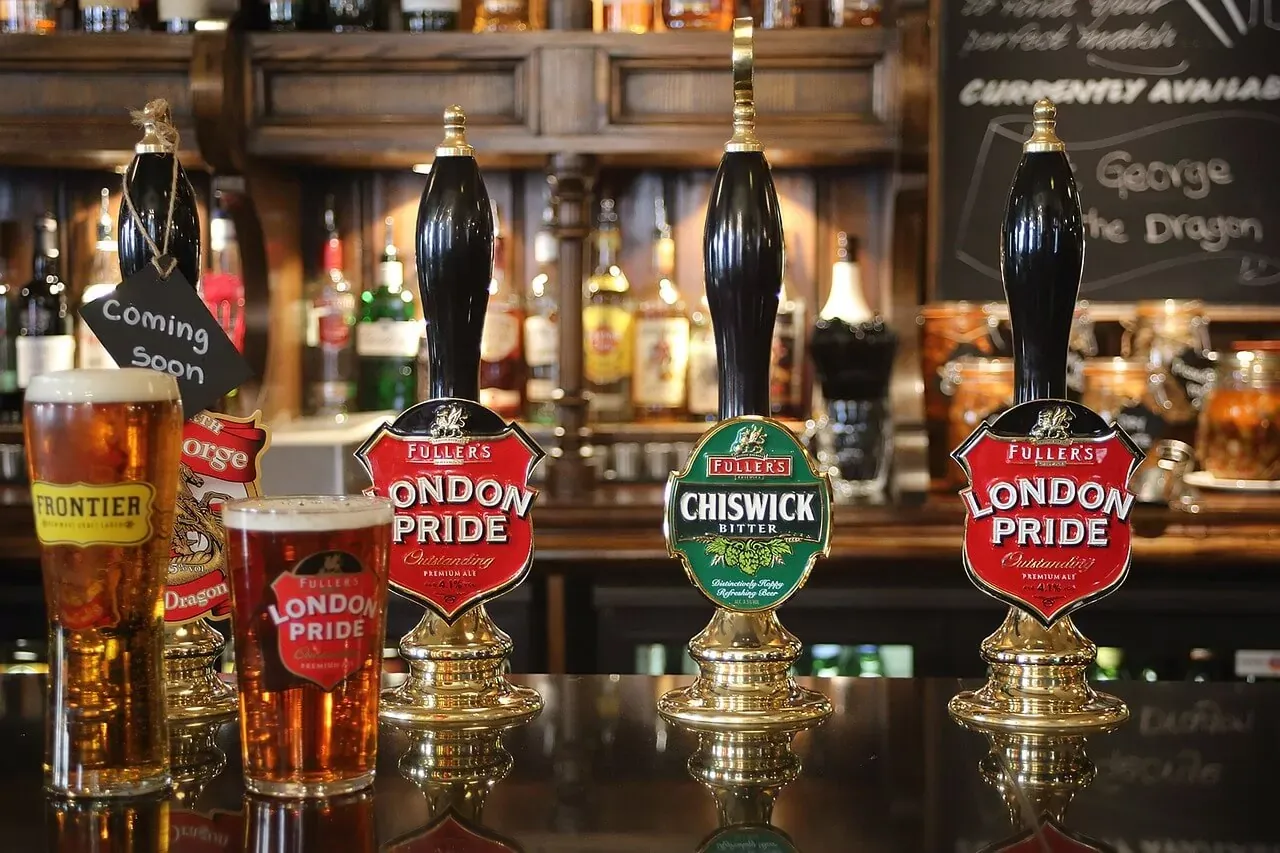 Cuatro grifos de cerveza Fuller's (London Pride, Chiswick Bitter) y dos pintas de cerveza en una barra de pub. Cartel 'Coming