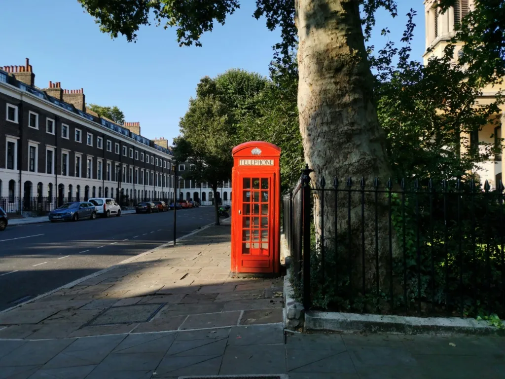 Klasyczna czerwona budka telefoniczna na słonecznej ulicy Londynu, w tle zabudowa szeregowa.