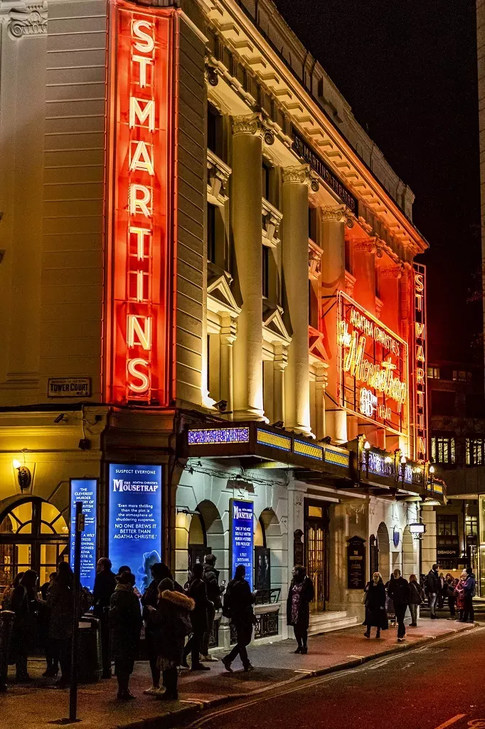 Exterior of St Martin's Theatre in London at night, with red neon signs, classical columns, and people on the pavement.