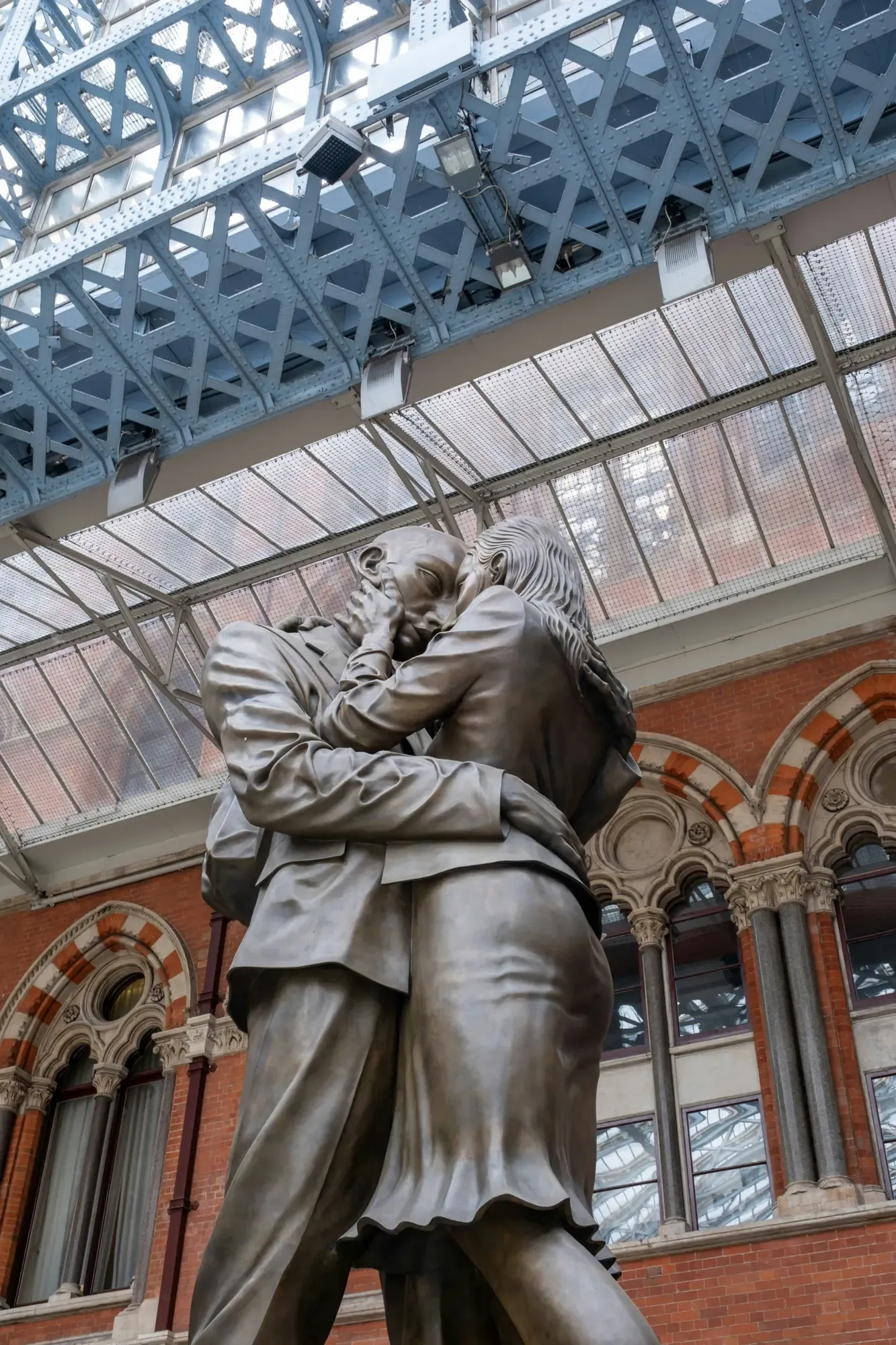 Eine riesige Bronzestatue eines Mannes und einer Frau, die sich unter dem gewölbten Glasdach des Bahnhofs St. Pancras umar...