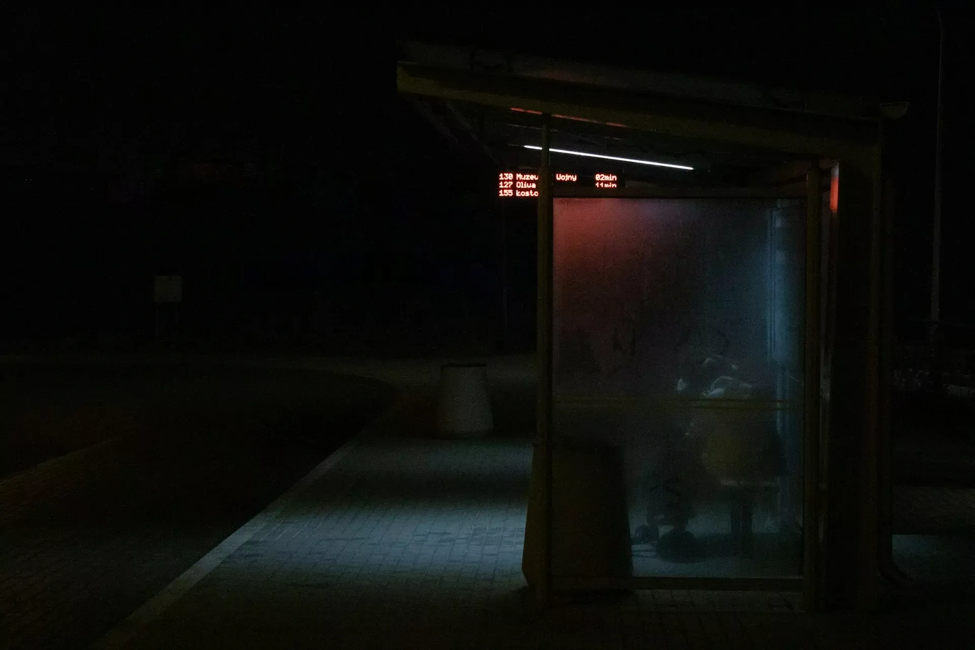 Bus stop at night with a digital display showing bus numbers, destinations like Oliwa, and arrival times in minutes.