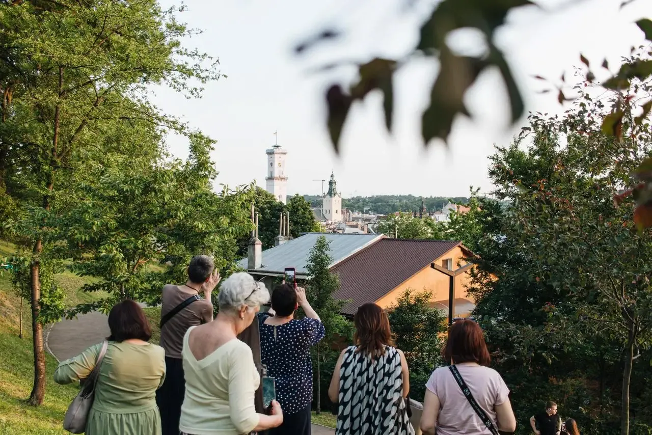 Grupa osób na wzgórzu podziwia panoramę Lwowa z widoczną wieżą Ratusza i kopułą kościoła wśród drzew.