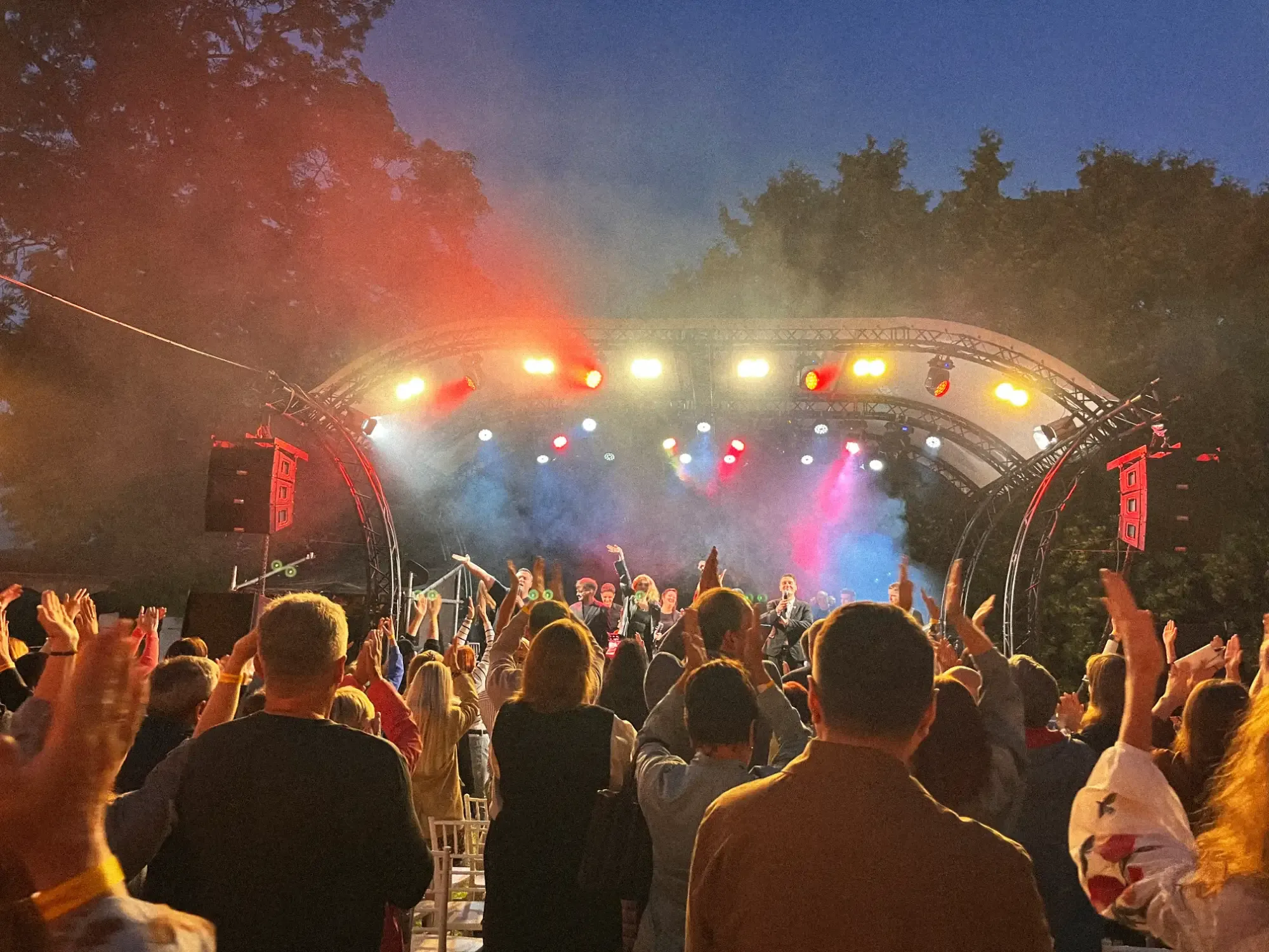 A crowd seen from behind watches performers on a brightly lit outdoor stage as dusk settles.
