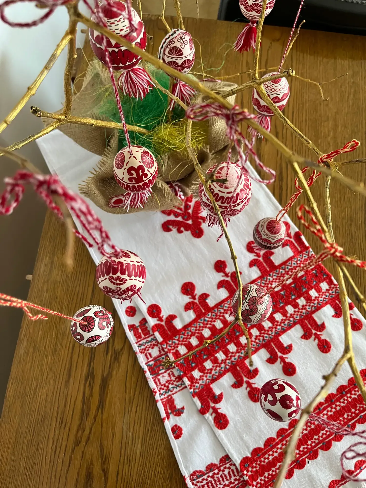 Traditional Ukrainian pysanky (Easter eggs) with red and white patterns hanging from golden branches over an embroidered tabl