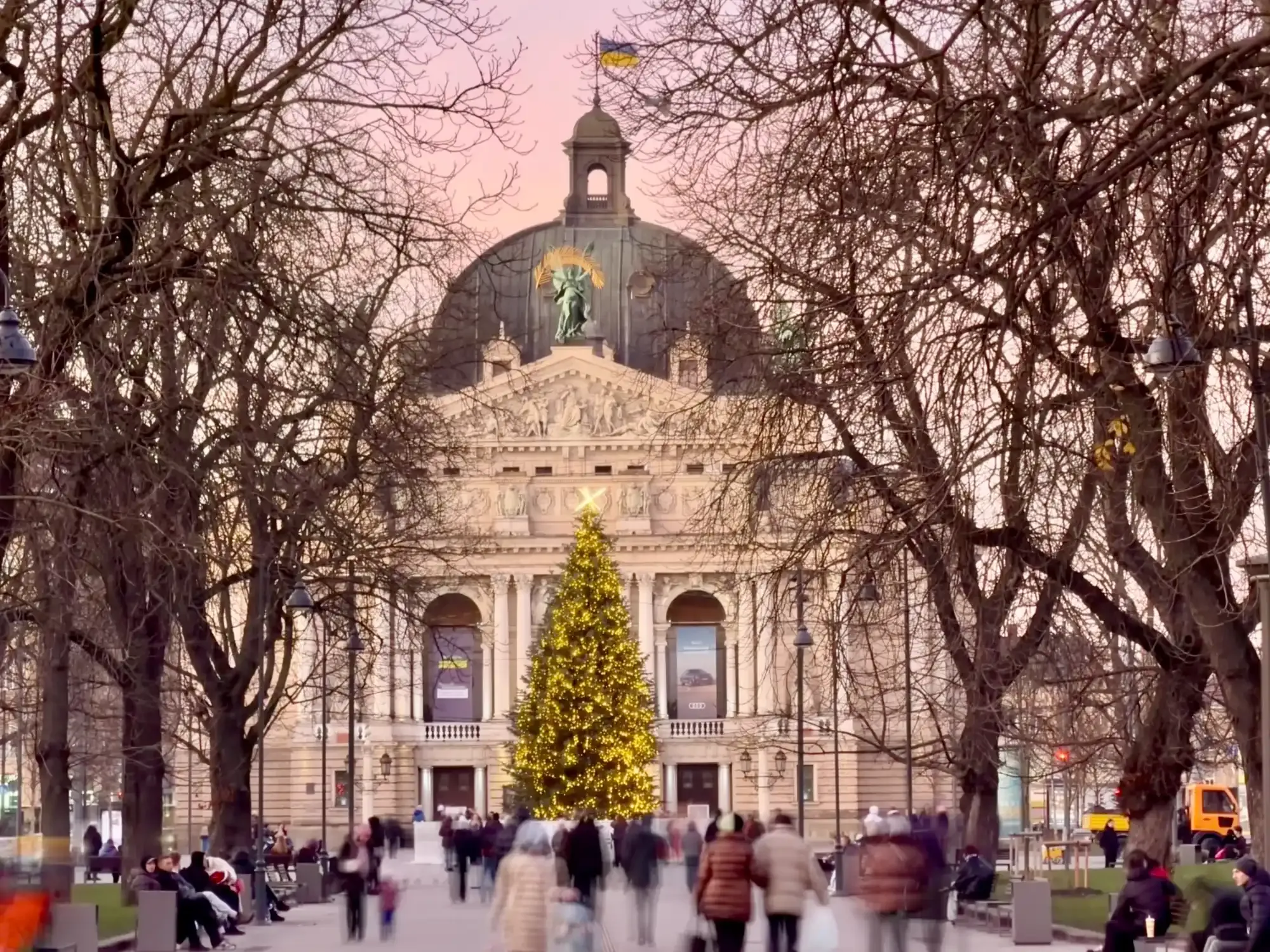 Grand classical building with a dome and Ukrainian flag, a large lit Christmas tree, and blurred people walking in front.