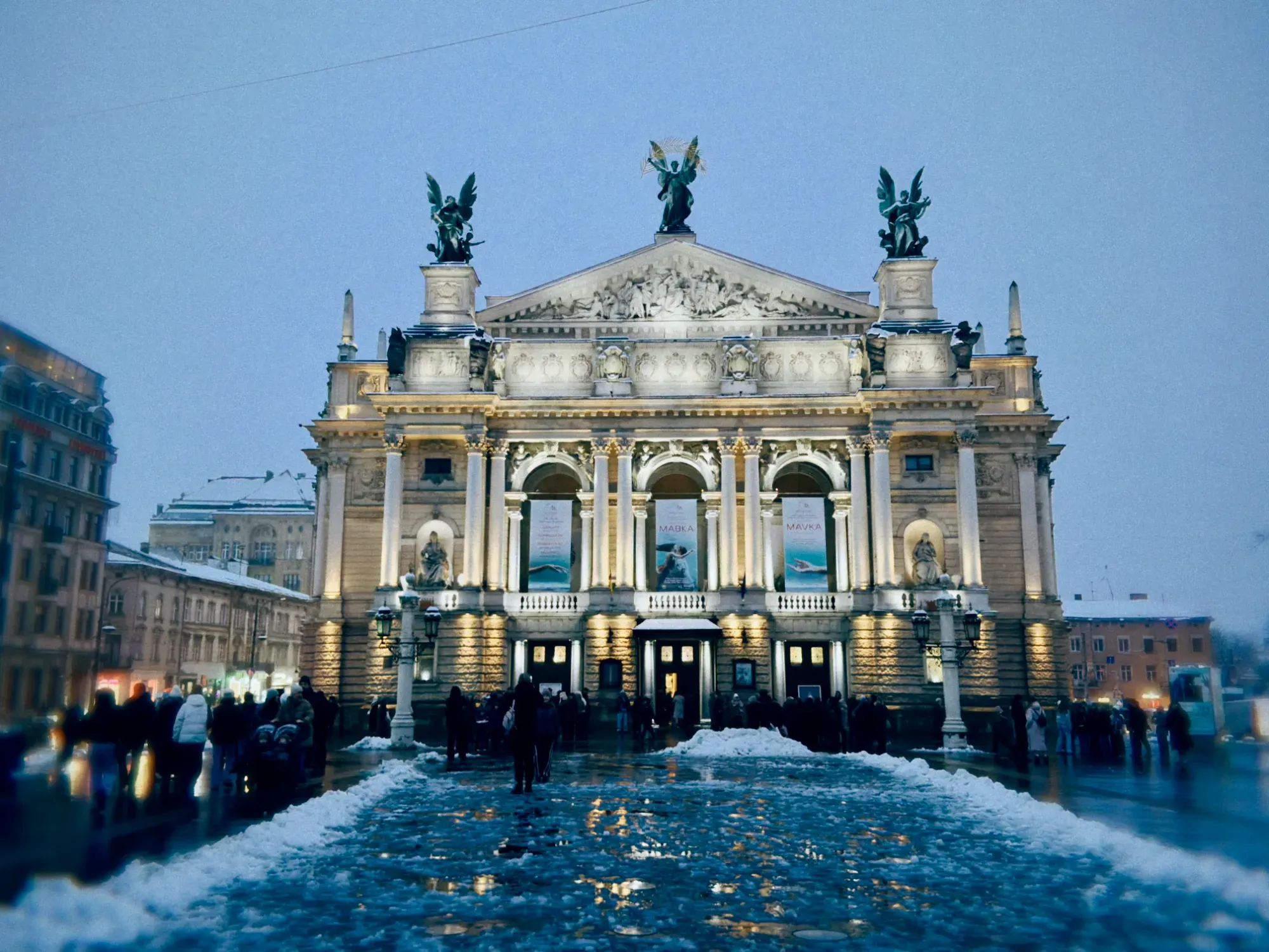 Lviv Opera House at dusk in winter, illuminated facade with statues, snow and slush on the ground, blurred people walking.