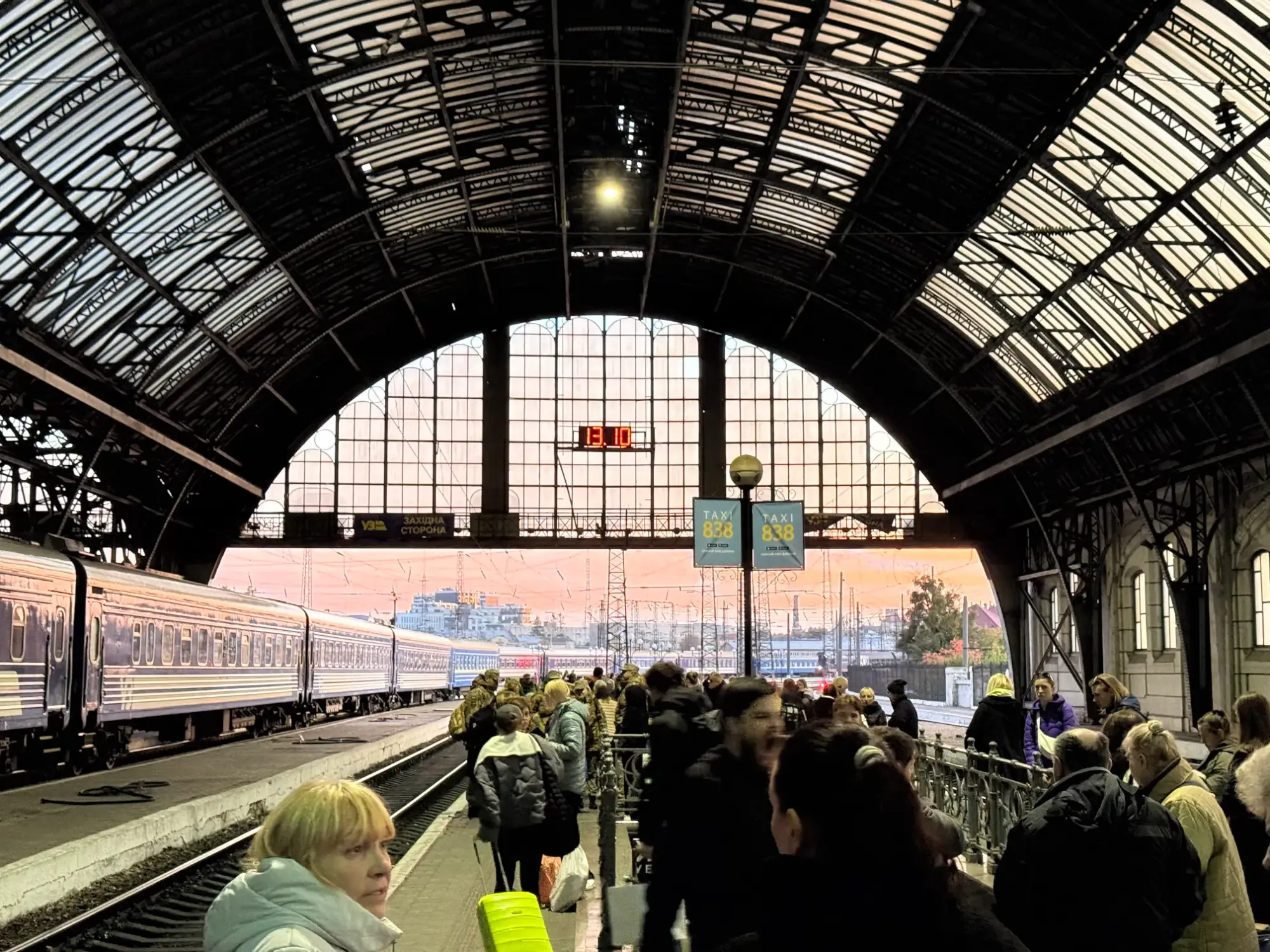 People wait on a train station platform next to a blue train under a large arched roof with a sunset sky.