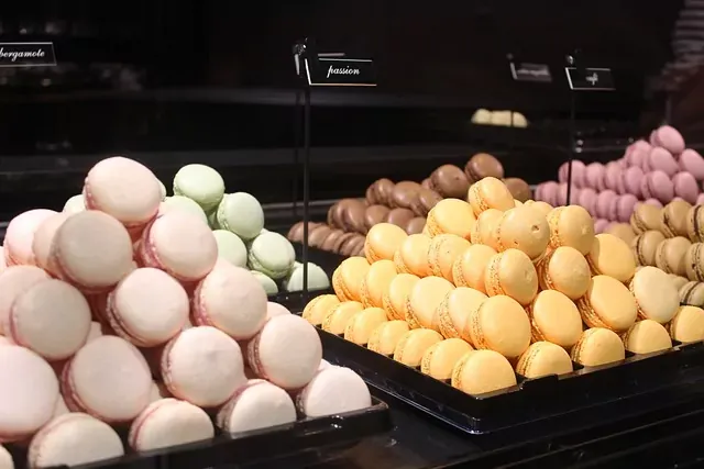 Haufen rosa, grüner, gelber, brauner und lila Macarons auf schwarzen Tabletts in einer Bäckerei-Auslage. Etiketten 'bergamote