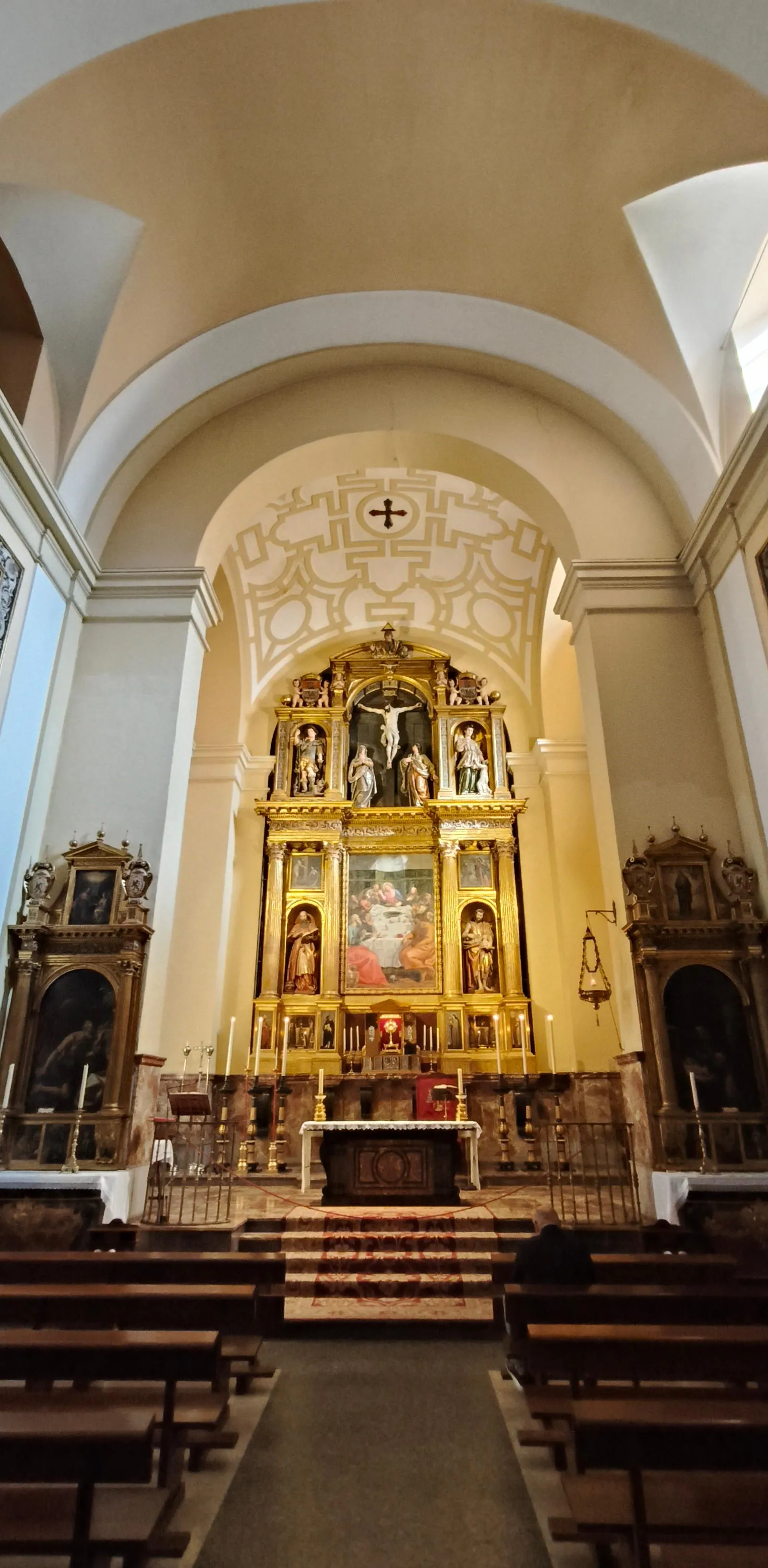 Ornate golden altarpiece inside a church with religious figures, paintings, and candles. Wooden pews are visible in the foreg