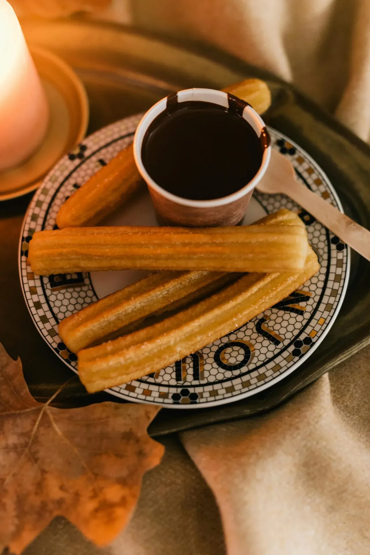 Teller mit mehreren gezuckerten Churros, einer Tasse dunkler Schokolade, einem Holzlöffel, einer brennenden Kerze und einem H