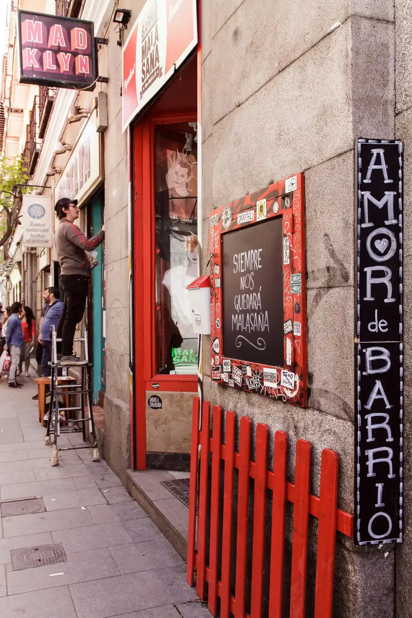 A street scene in the Malasaña neighborhood of Madrid, with signs, shops, and a man standing on a ladder.