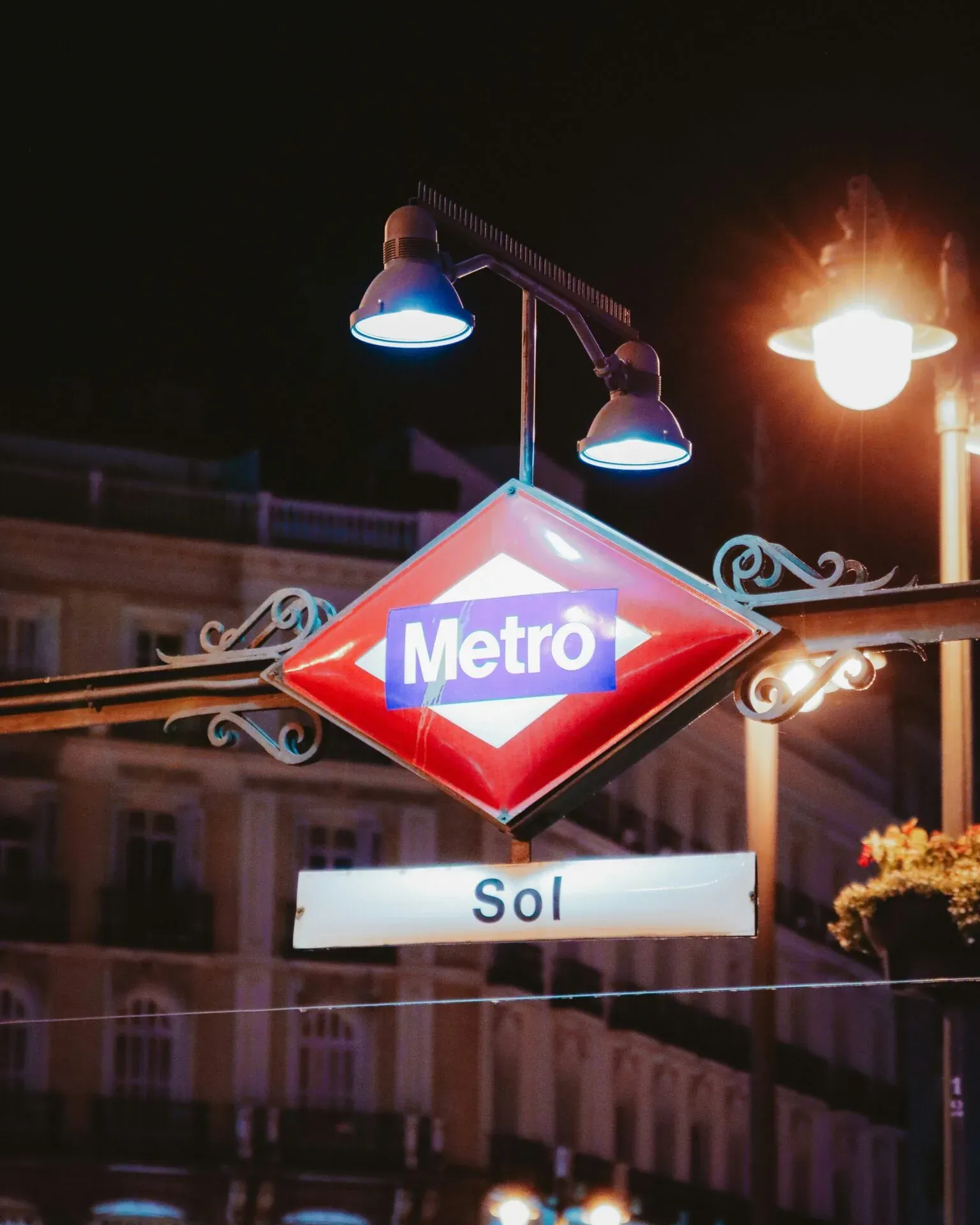 Illuminated red diamond Madrid Metro sign for Sol station at night, with streetlights and building facades in the blurred bac