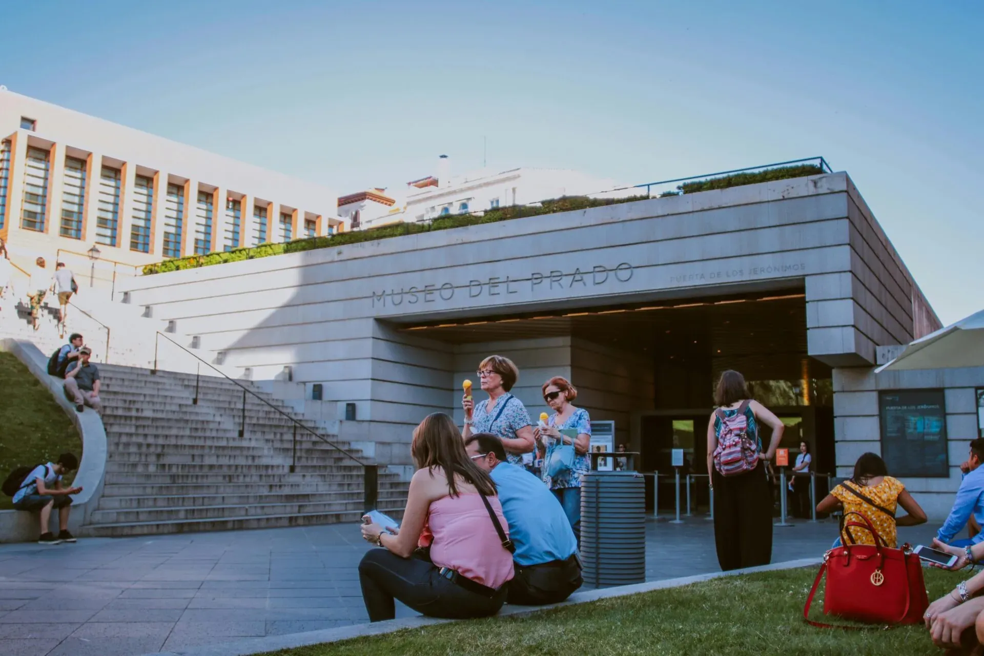 People relaxing and eating ice cream outside the modern entrance of the Museo del Prado in Madrid. The building shows 'MUSEO