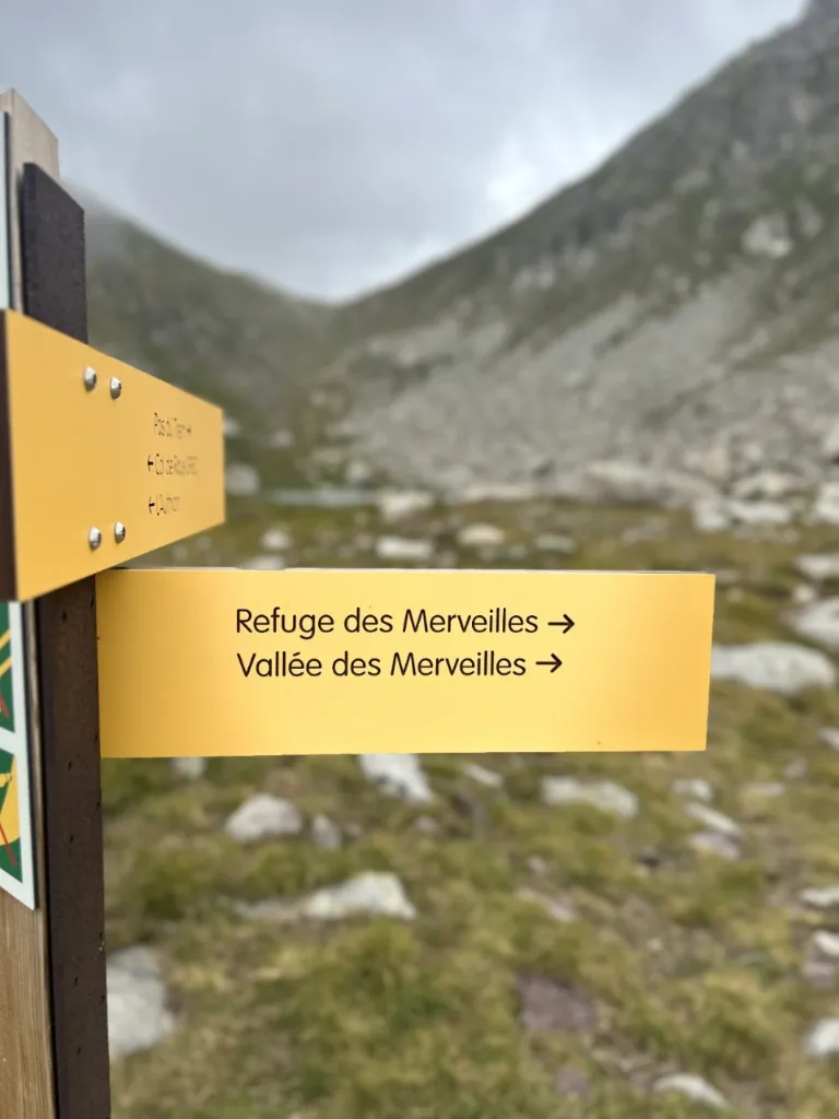 Gelber Wanderwegweiser in felsiger Berglandschaft unter bewölktem Himmel, zeigt auf 'Refuge des Merveilles' und 'Vallée des M