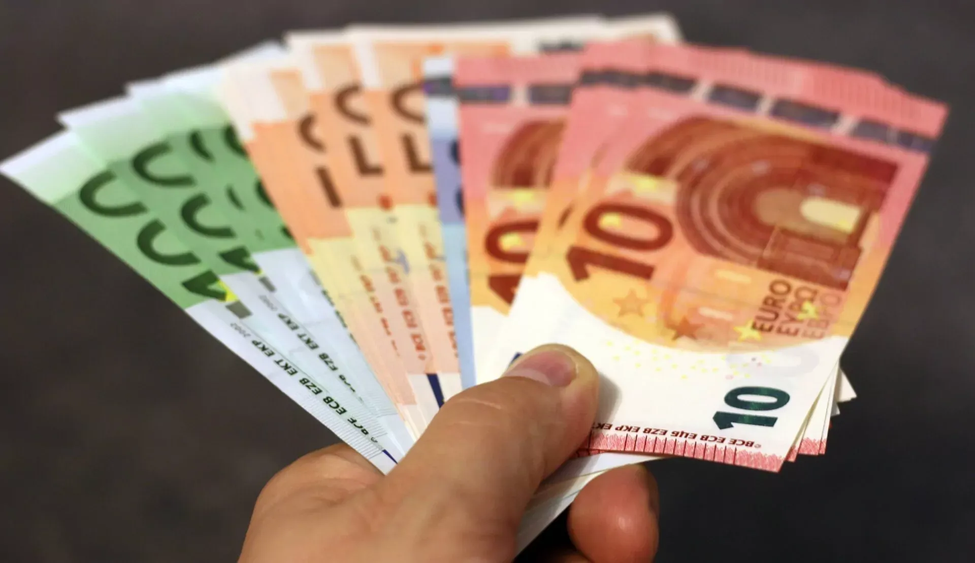 A hand holds a fan of various Euro banknotes, including 10, 20, 50, and 100 denominations, against a dark background.