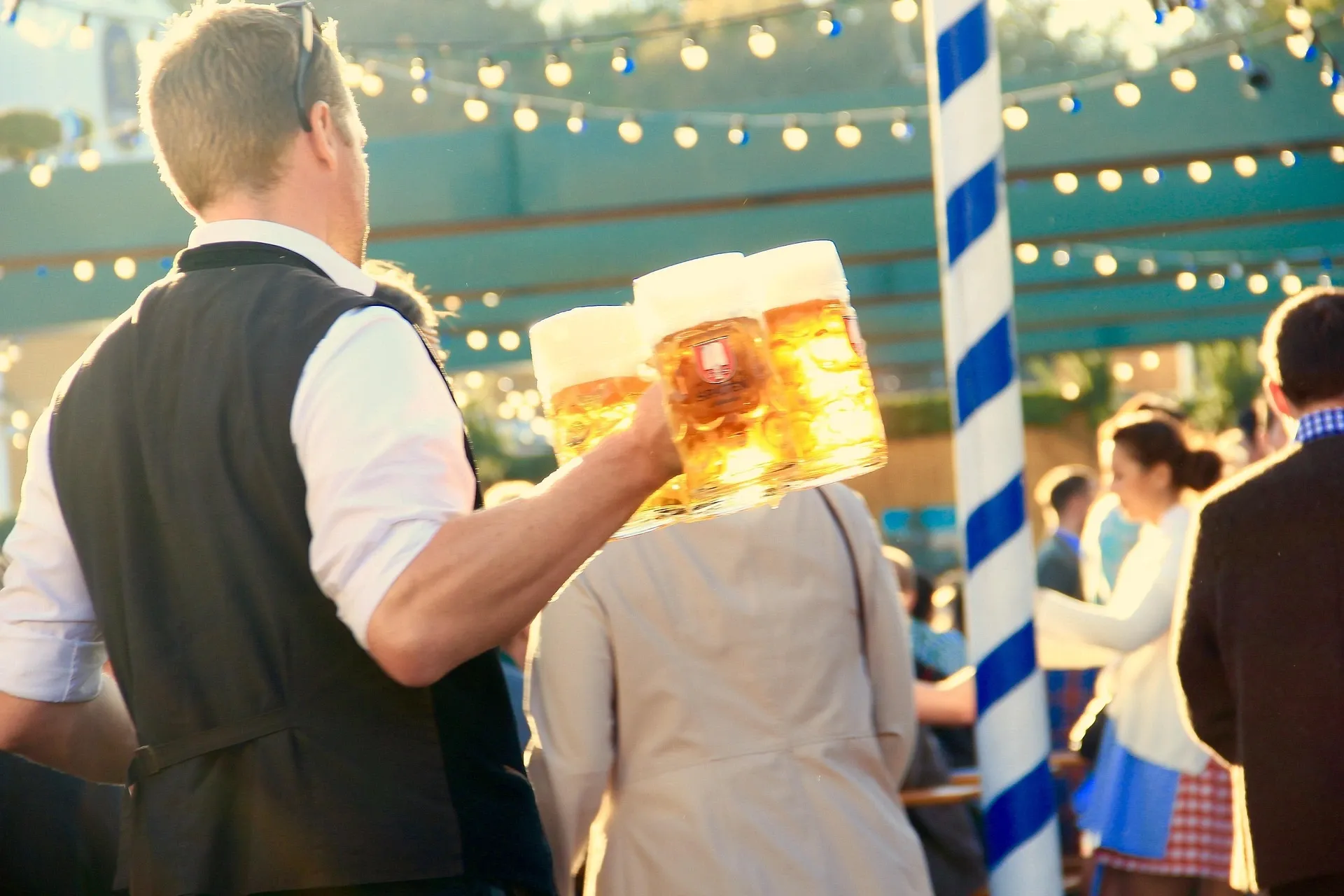 A person carries three large steins of frothy beer. In the background are blurry people, string lights, and a blue and white