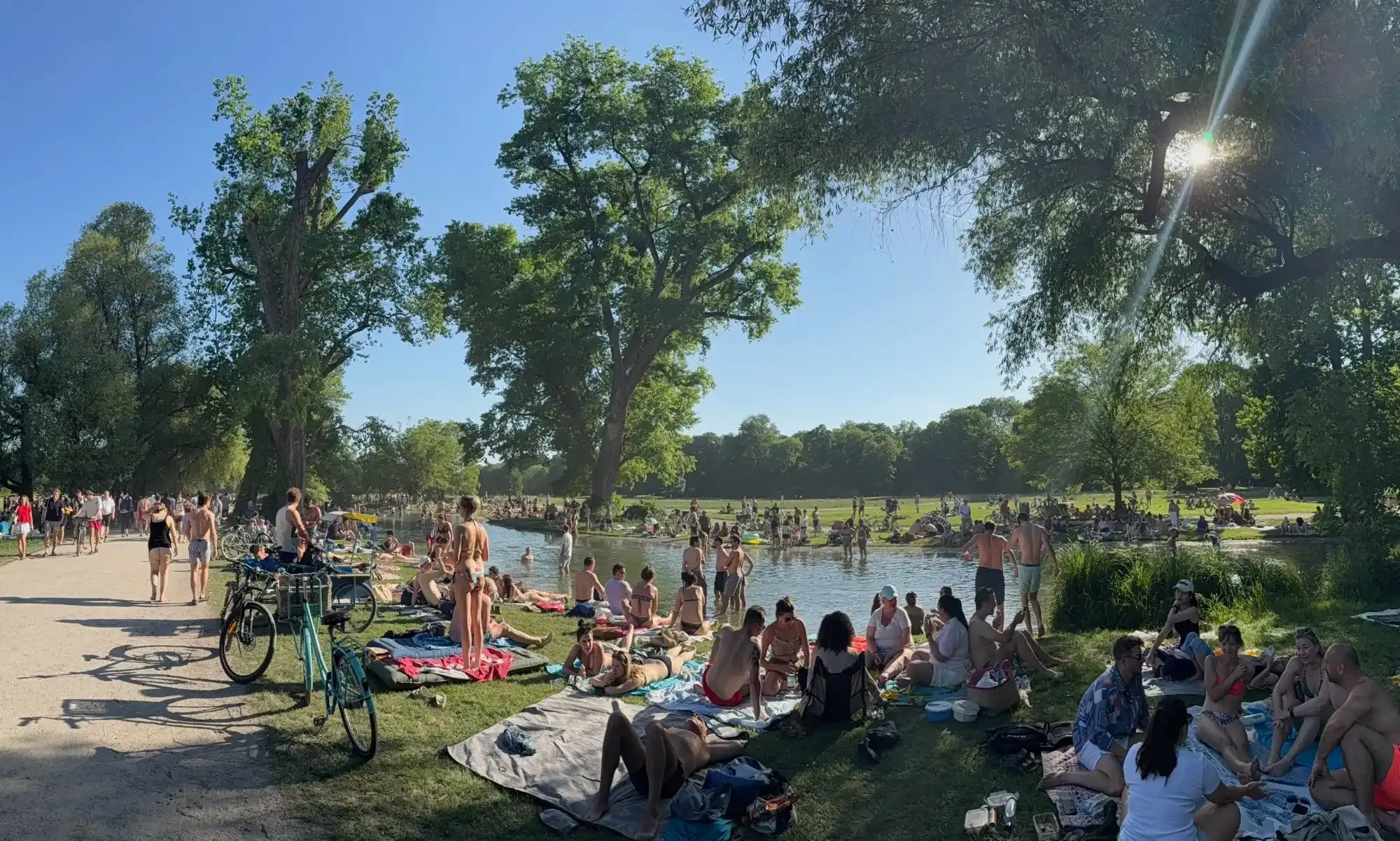 Crowds of people sunbathing and swimming in a river that runs through a green park in Munich on a sunny day.