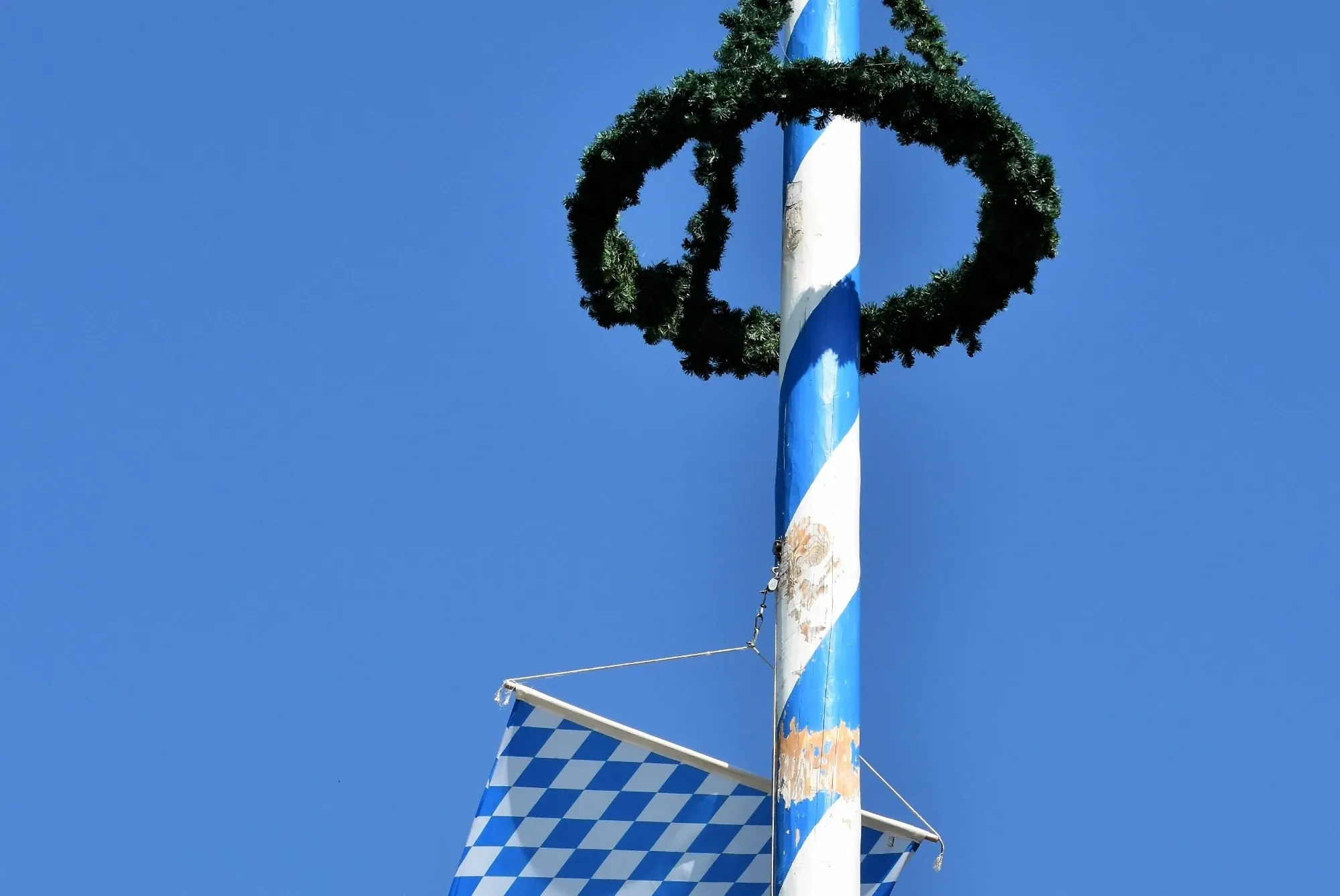 Blau-weißer Maibaum mit grünem Kranz und bayerischer Flagge vor klarem Himmel