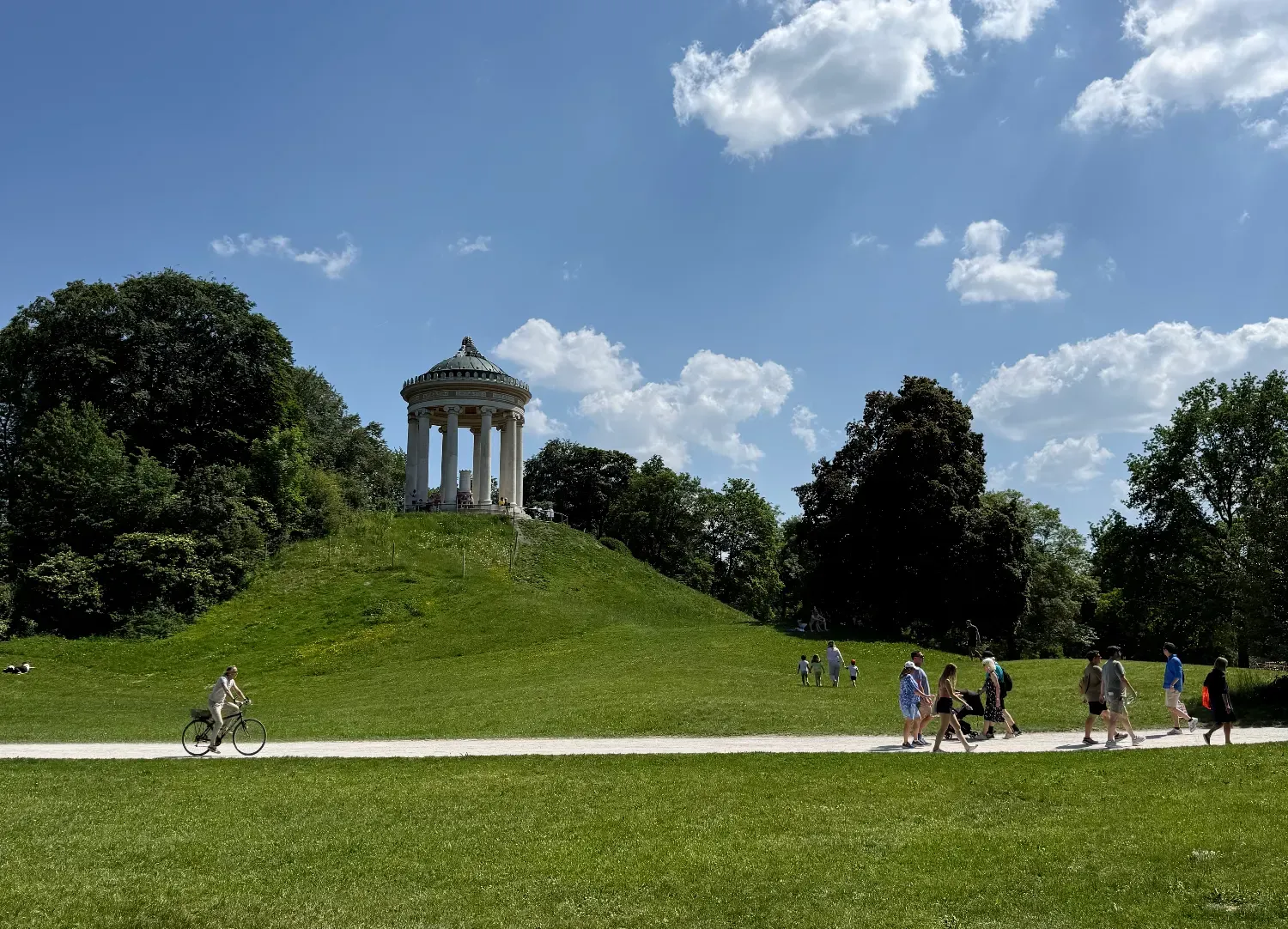 Der Monopteros-Tempel auf einem grünen Hügel im Englischen Garten, mit Menschen, die auf dem Weg spazieren gehen.
