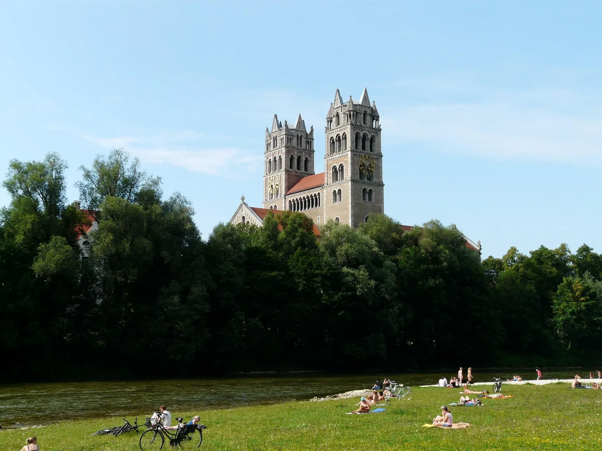 St. Maximilian Church with two towers and red roof, seen over trees from the grassy bank of the Isar River, where people rela