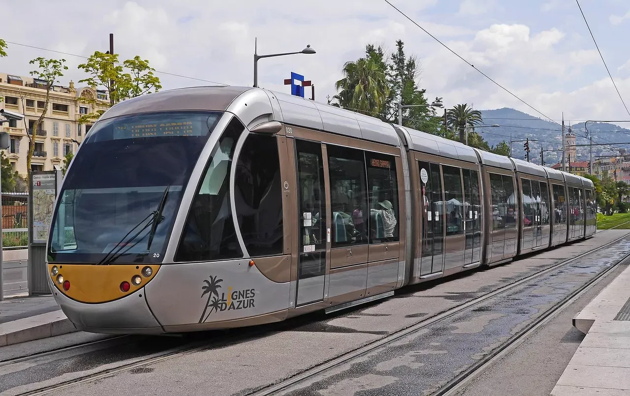 Nowoczesny srebrno-brązowy tramwaj z logo "Lignes d'Azur" na torach ulicznych w Nicei. W tle widoczne budynki i palmy pod zac