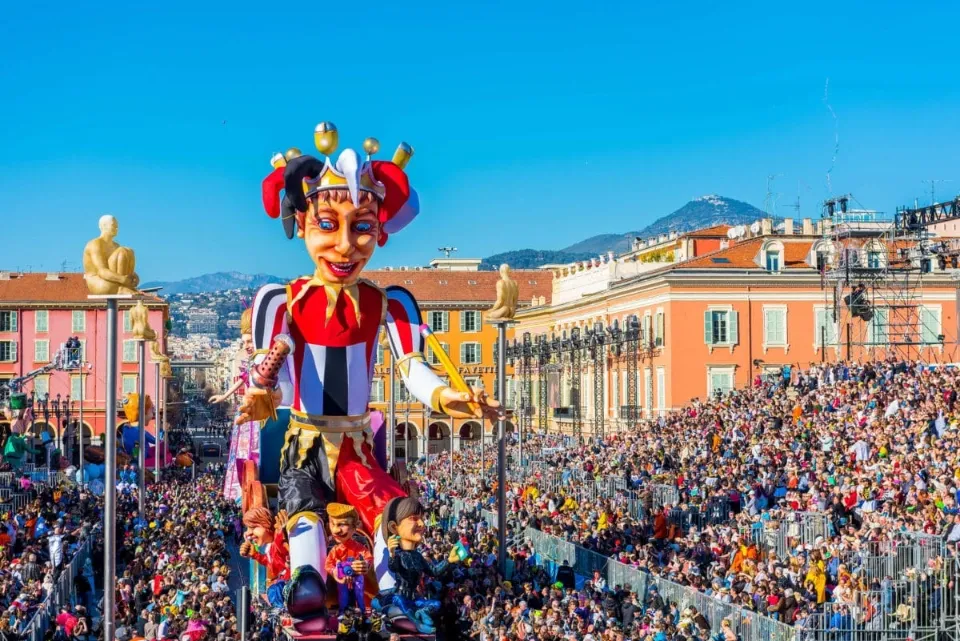 Gran carroza de carnaval con un bufón gigante y una multitud de espectadores en una plaza soleada. Edificios y montañas al fo
