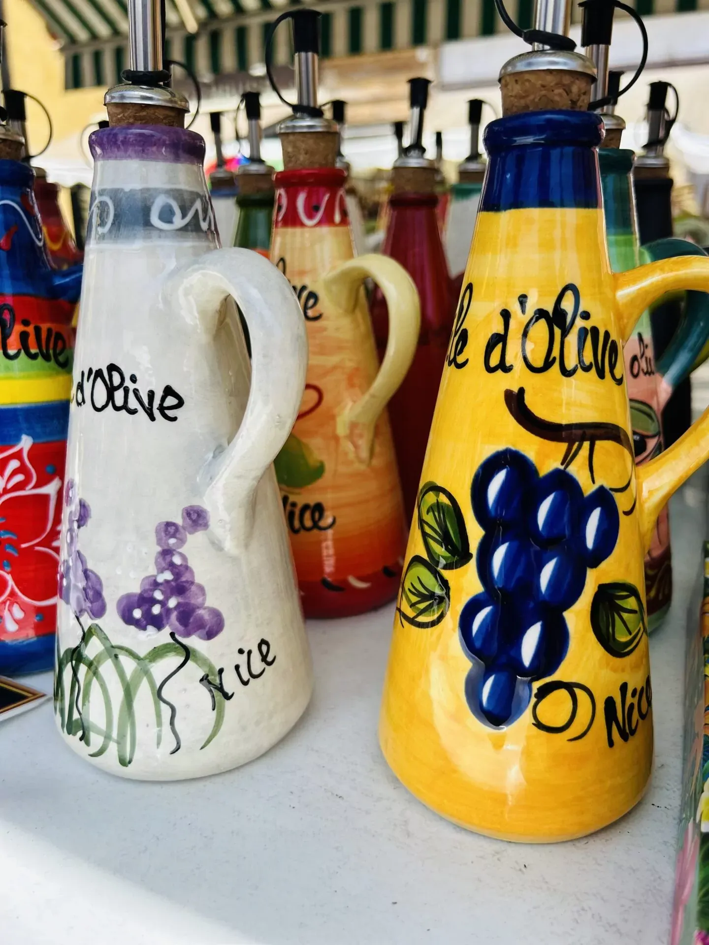 Close-up of colorful ceramic olive oil dispenser bottles with cork and metal spouts, some decorated with grapes and flowers,