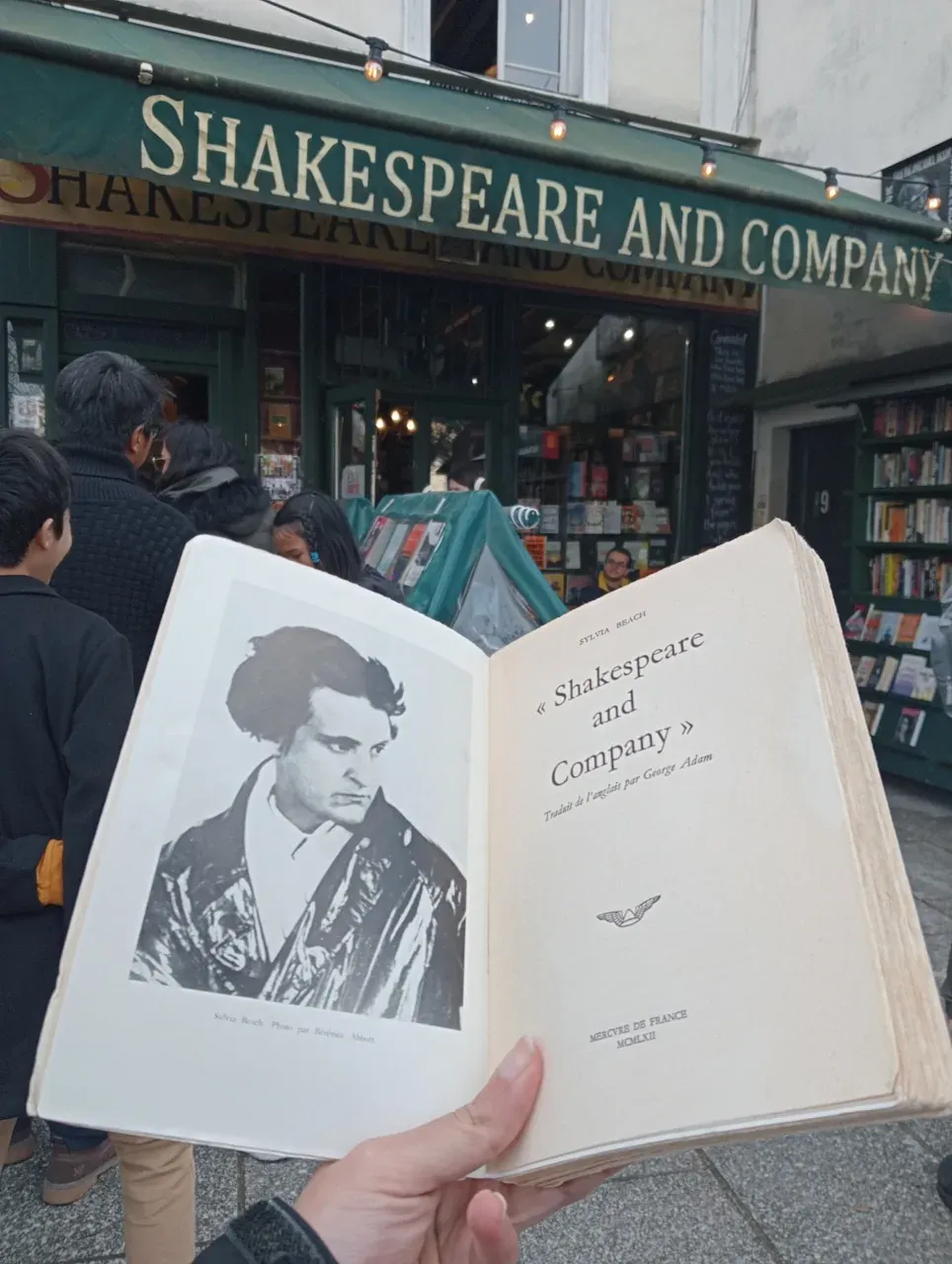 Dłoń trzymająca otwartą książkę ze zdjęciem Sylvii Beach przed księgarnią Shakespeare and Company w Paryżu.