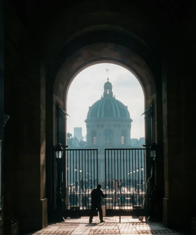 The domed Institut de France seen through a dark archway and iron gates in Paris, with silhouettes of pedestrians.