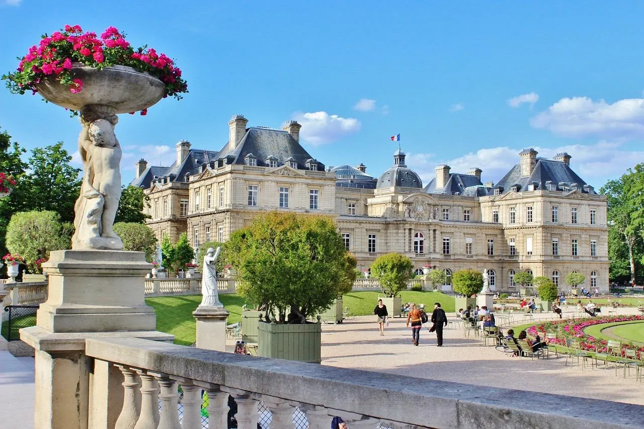 Vista del Palacio de Luxemburgo desde los jardines, con balaustrada de piedra, estatuas, flores y gente paseando.