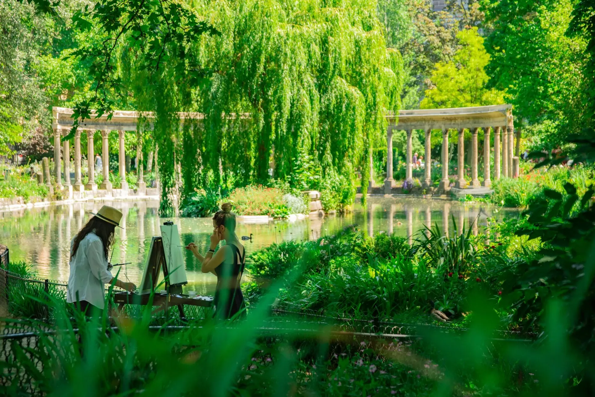 Two artists painting by a lake in a lush green park with a classical colonnade in the background in Paris.