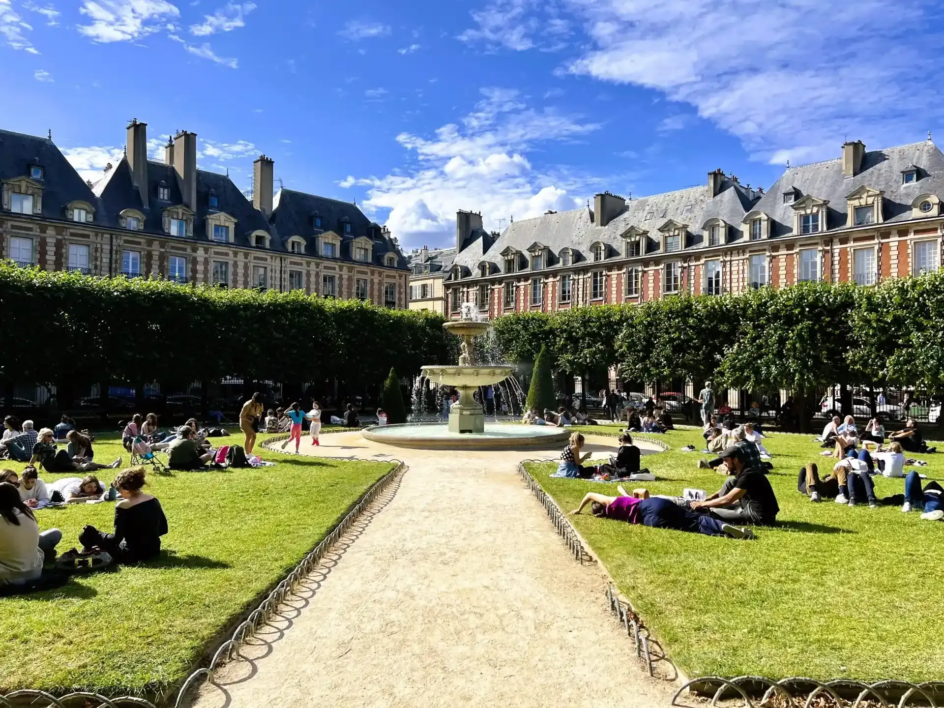 Place des Vosges en París con una fuente central, césped verde, gente relajándose y edificios históricos de ladrillo bajo cie