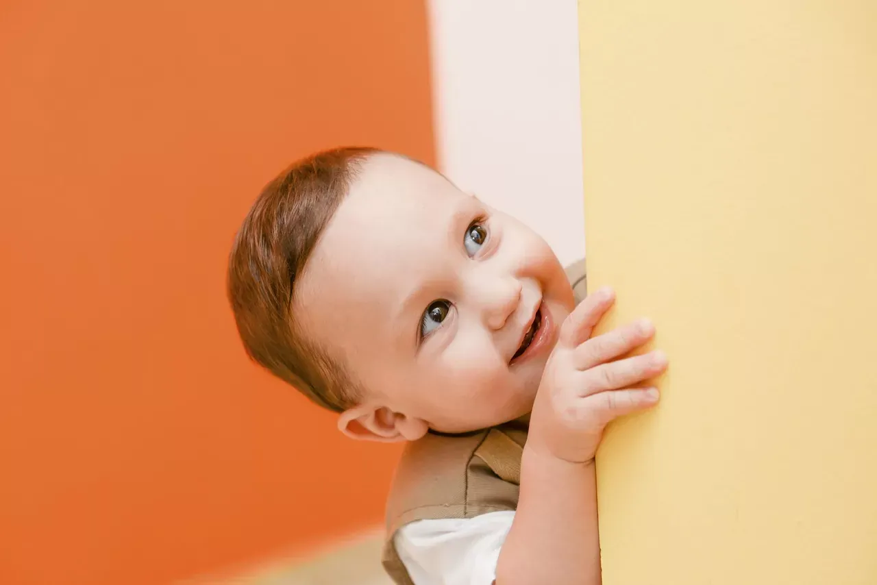 Happy baby peeking from behind a yellow wall, smiling with brown eyes, against an orange background.