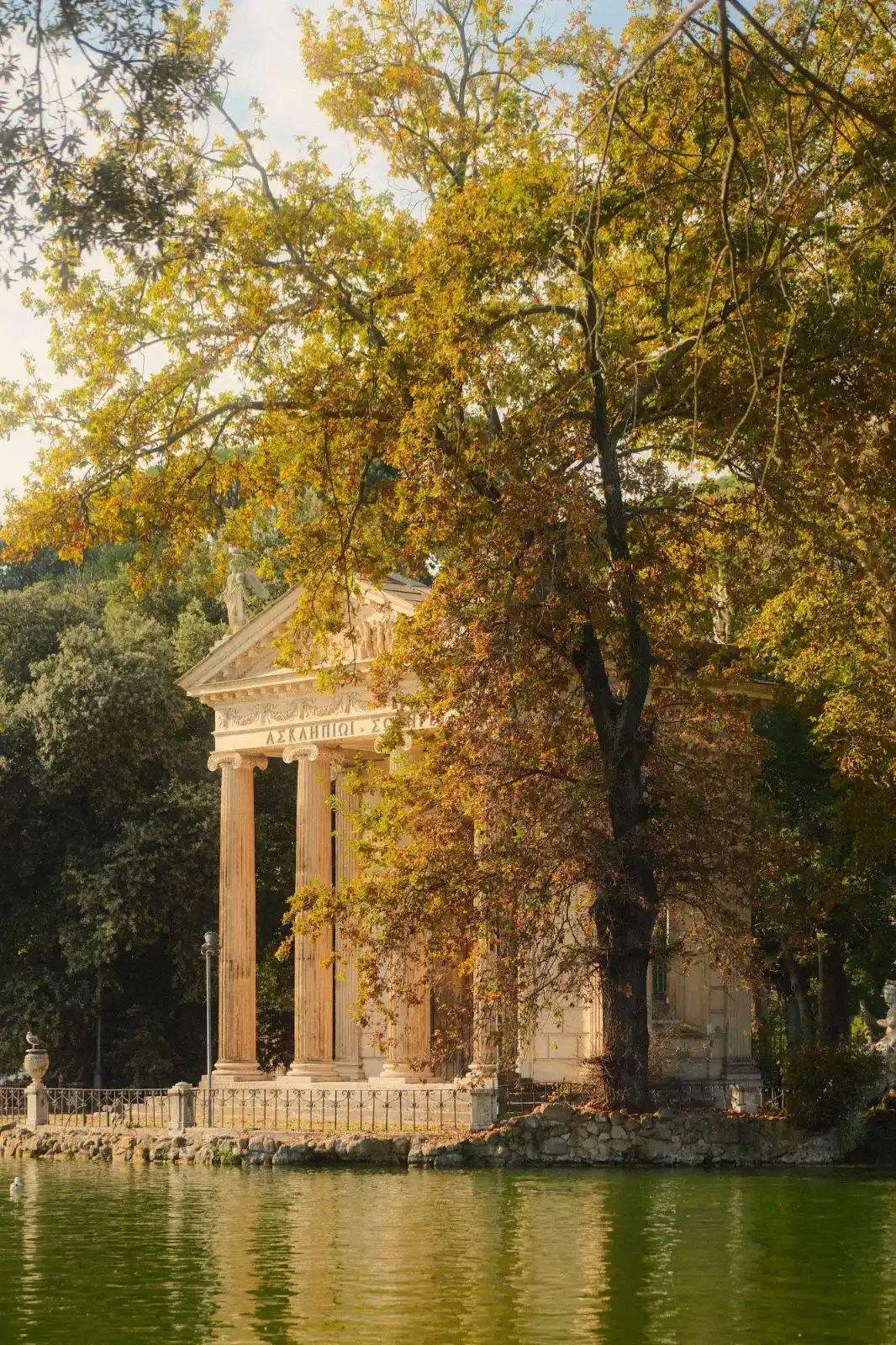 The Temple of Asclepius, a classical-style structure with columns, seen across a lake in the Borghese Gardens in autumn.