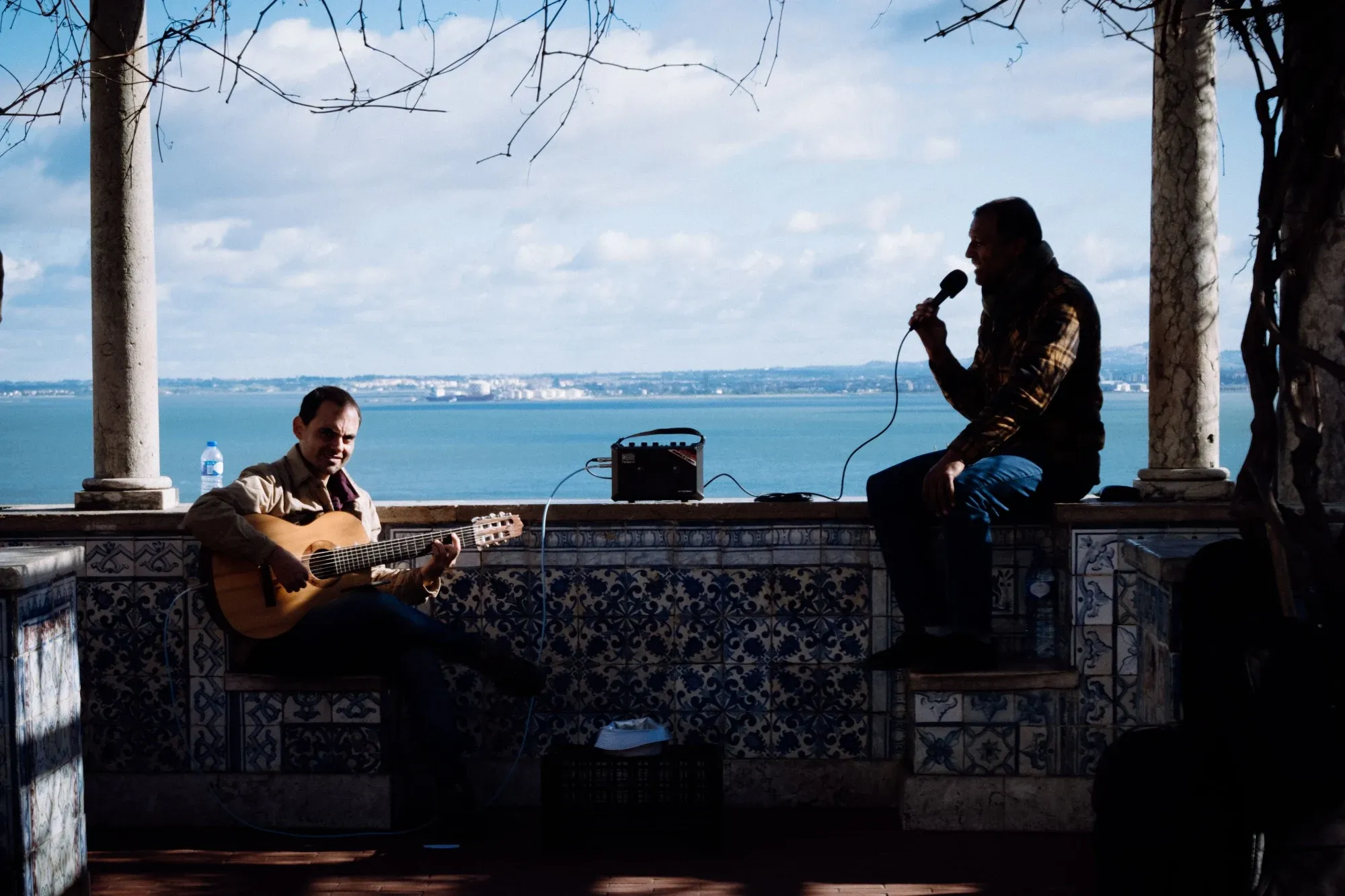 Two musicians perform with guitar and microphone at a tiled viewpoint overlooking the Tagus River in Lisbon.