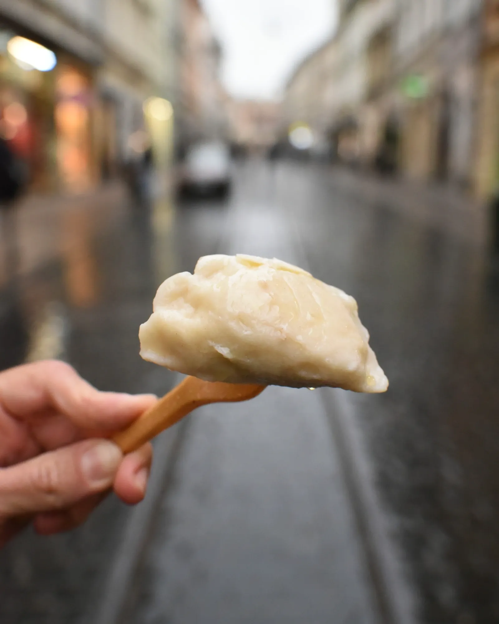 Hand holding a traditional Polish pierogi dumpling on a wooden fork against a blurred Krakow street background.