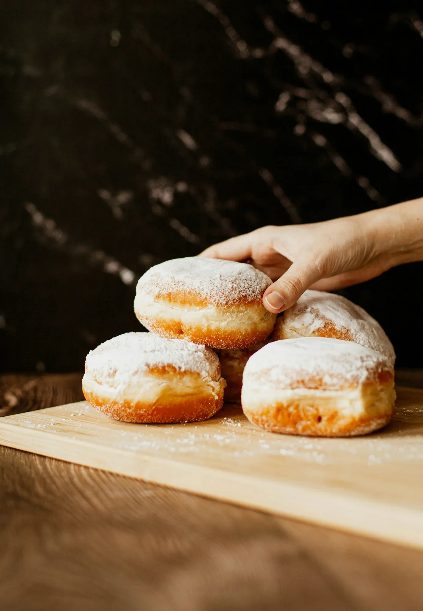 A hand reaches for a sugar-dusted pączek from a pile of several traditional Polish doughnuts on a wooden board.