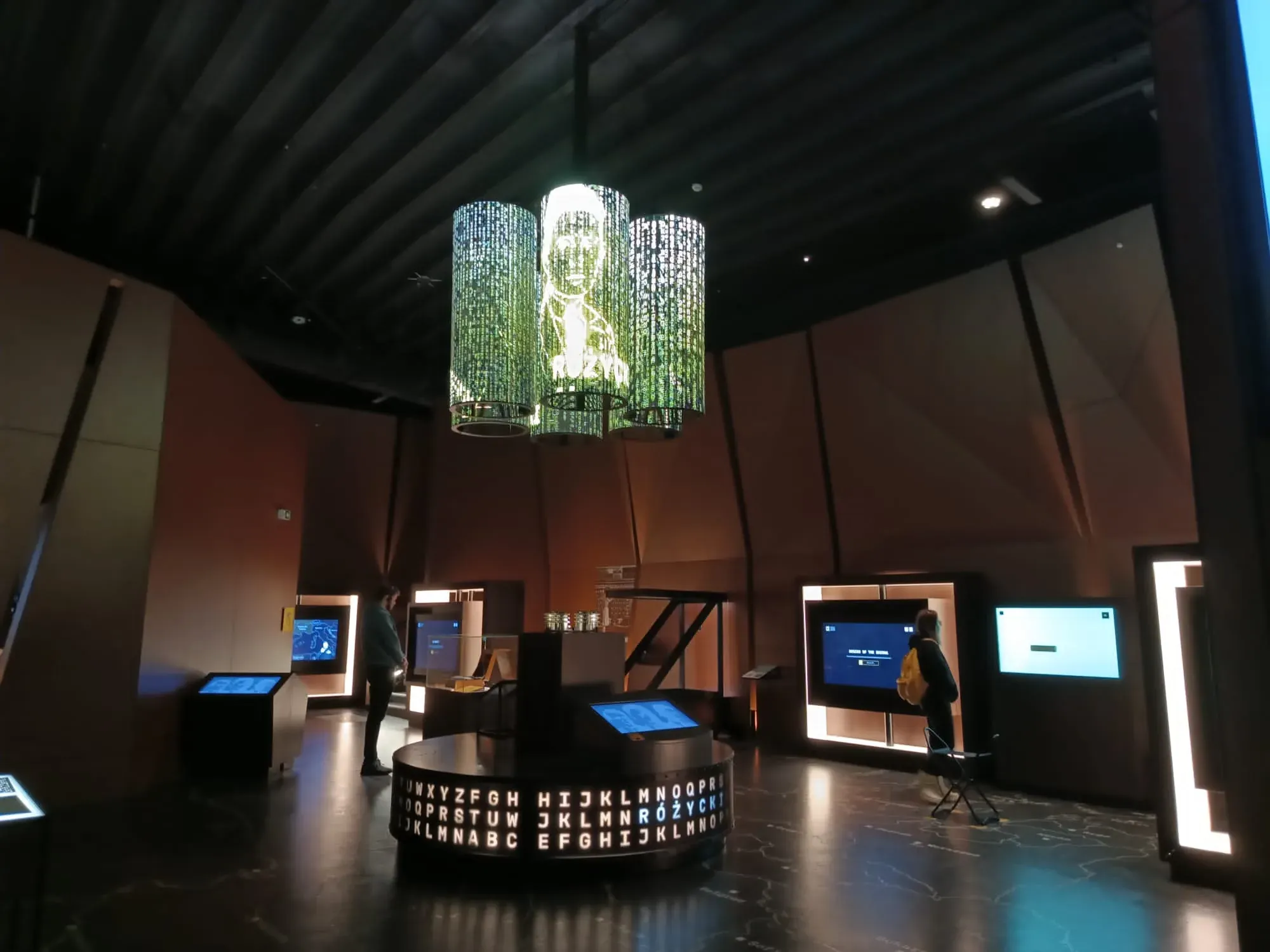 Interior moderno de museo con exposiciones interactivas, visitantes y una gran pantalla redonda de letras en el suelo.