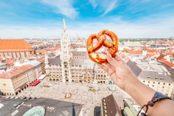 Eine Hand hält eine Brezel über den sonnigen Marienplatz in München, mit dem Neuen Rathaus im Hintergrund.