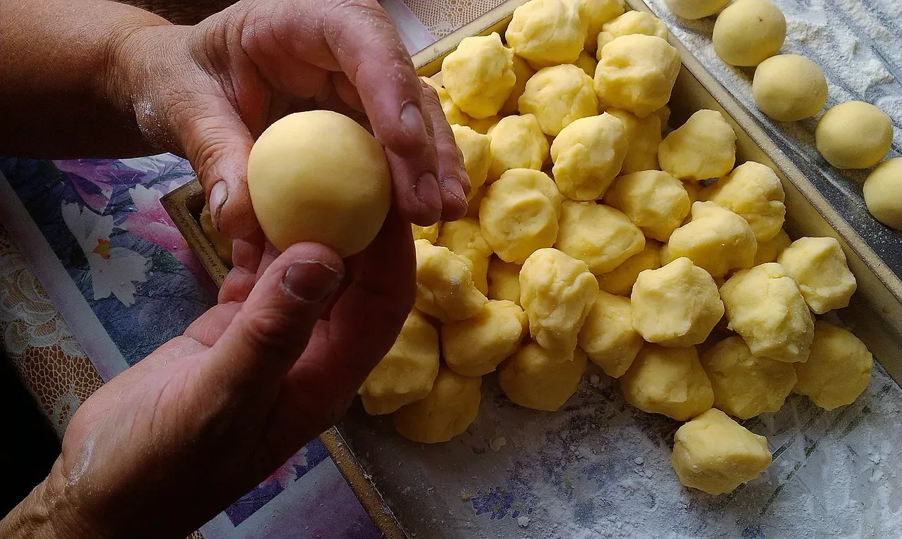 Manos sosteniendo una bola de masa de patata redonda y amarilla, con una bandeja de más trozos de masa enharinados.