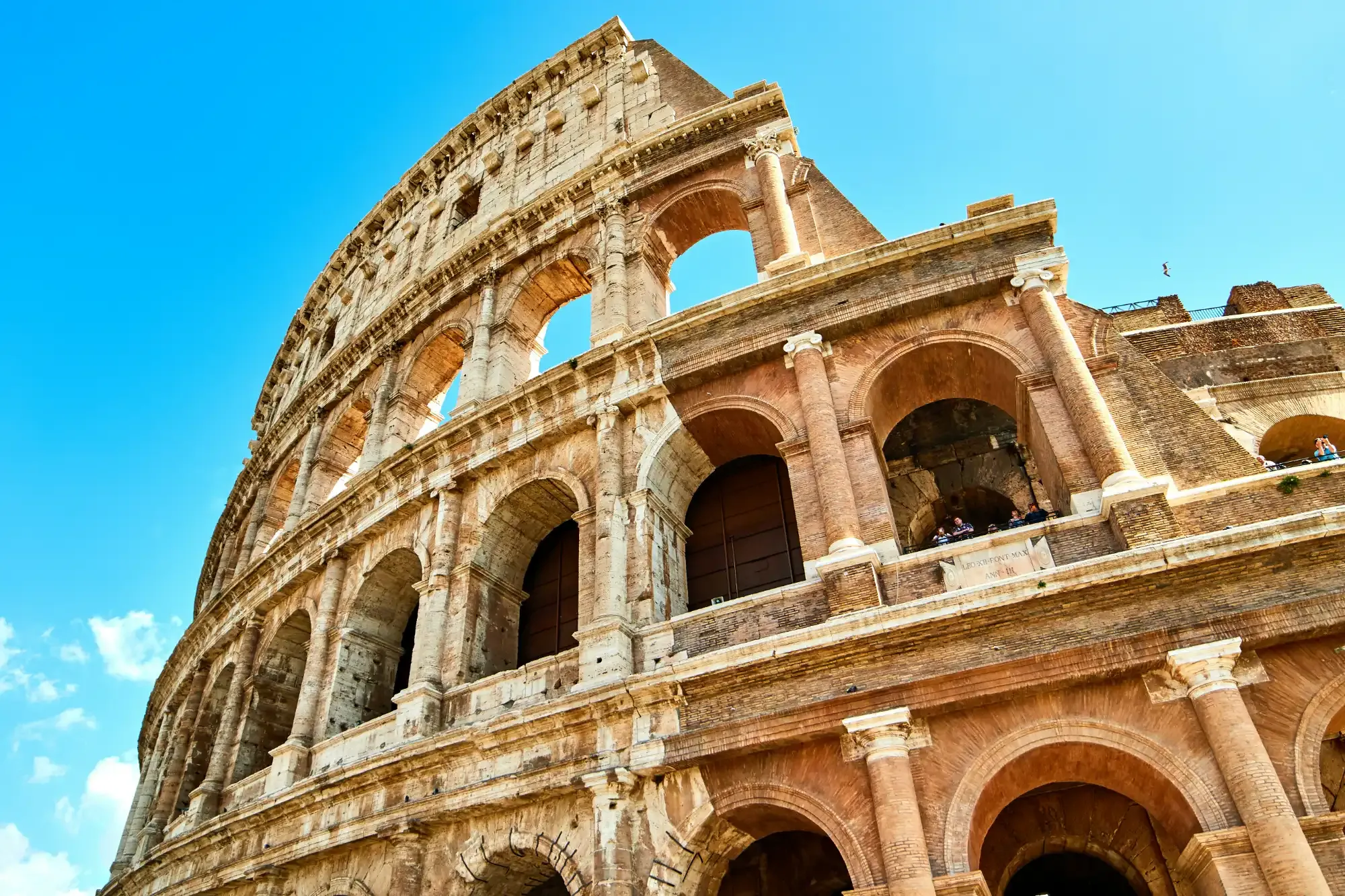Low-angle view of the Colosseum's exterior stone arches in Rome under a bright blue sky.