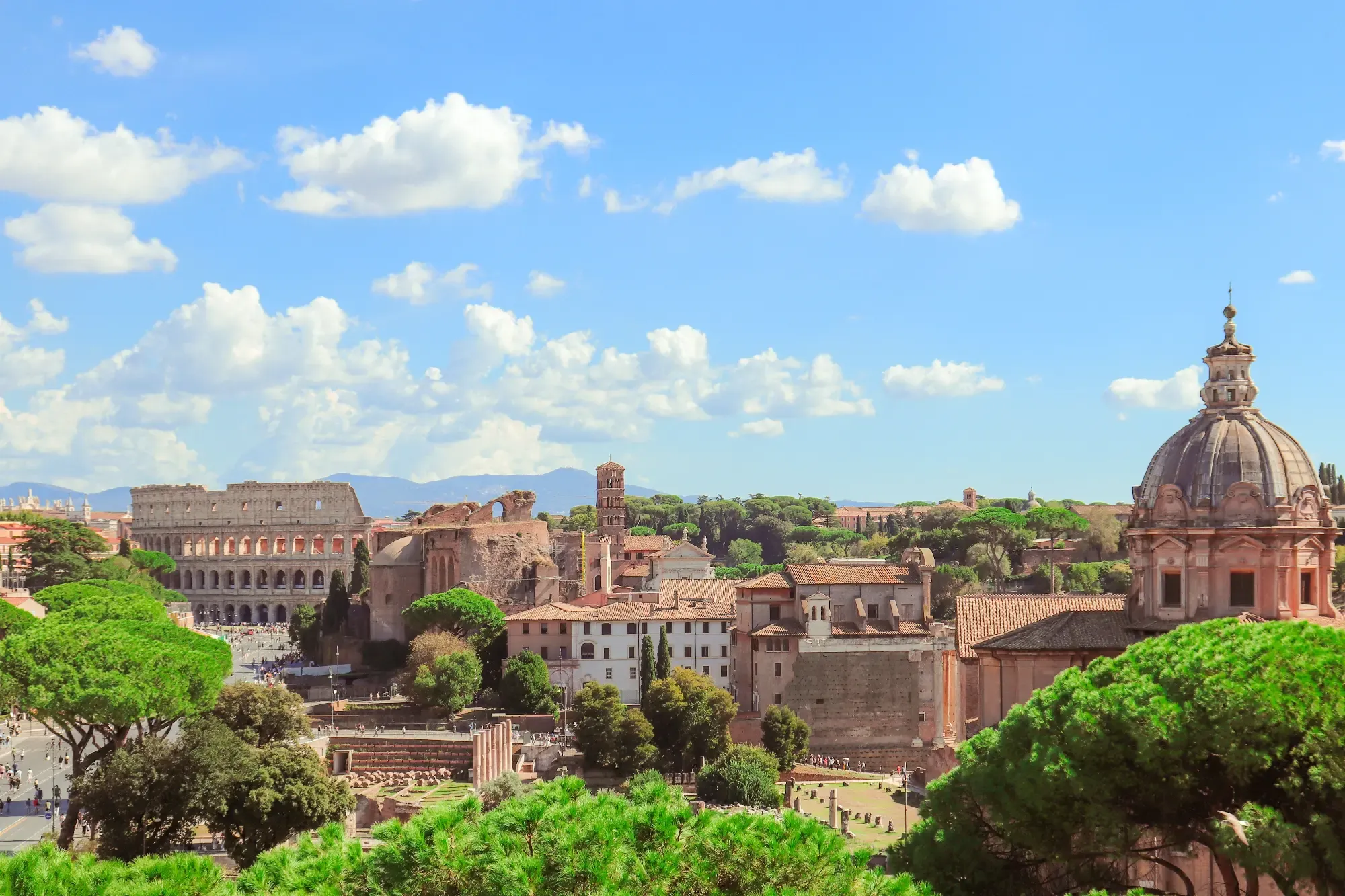 View of the Roman Forum and the Colosseum in Rome under a blue sky with clouds, seen from a high vantage point.