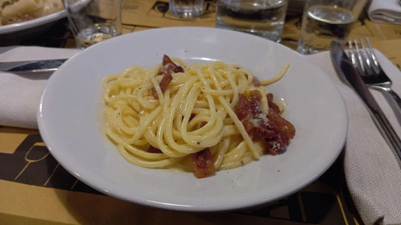 Plato de espagueti carbonara con guanciale, queso rallado y pimienta negra sobre una mesa de restaurante con cubiertos y vaso