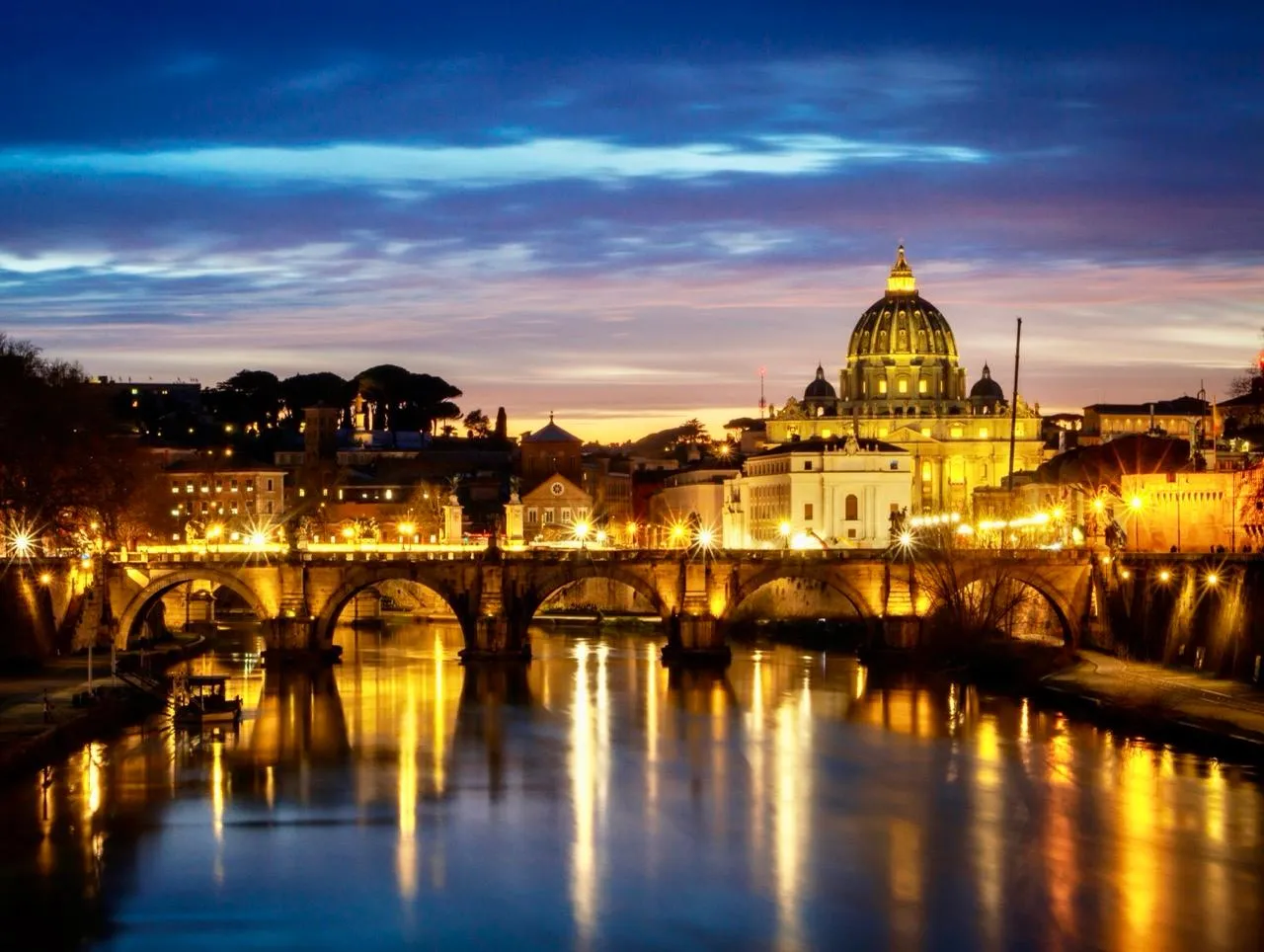 St. Peter's Basilica and Ponte Sant'Angelo over the Tiber River at twilight, with city lights reflecting on the water.